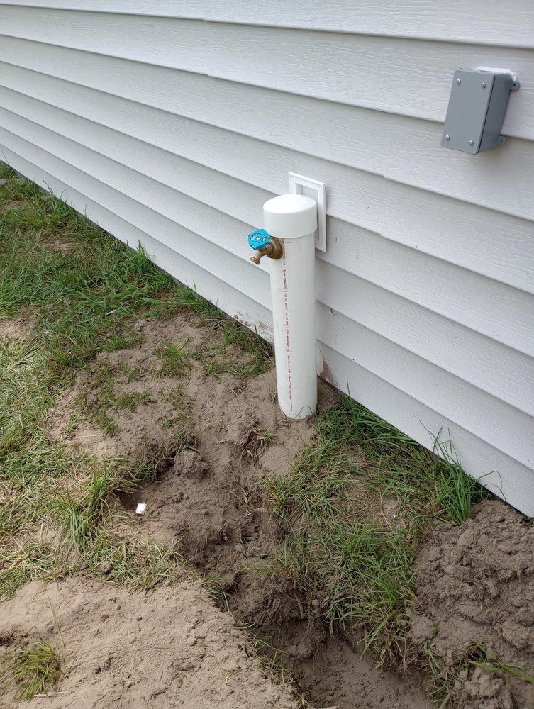A white PVC pipe housing an outdoor water spigot against a house with light grey vinyl siding.