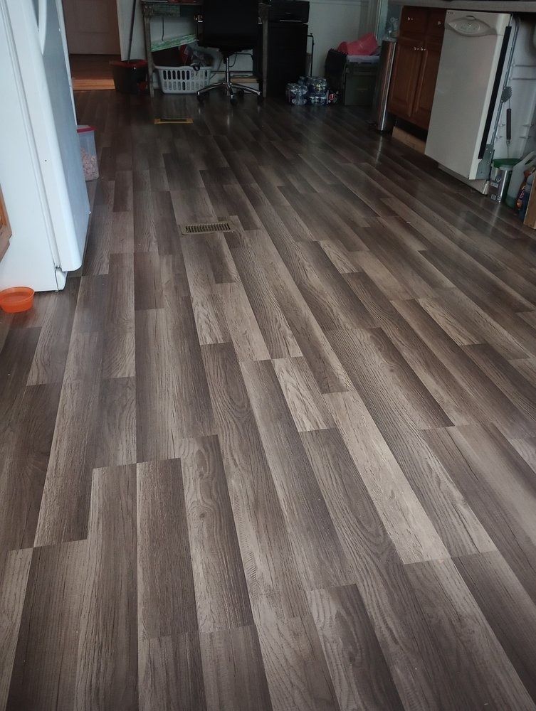 A wide-angle view of a residential floor featuring gray-toned wood-look laminate planks, leading toward a kitchen area.