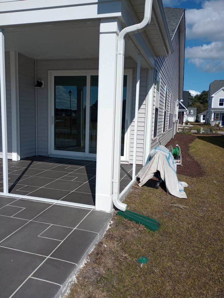 A white downspout on a house exterior directs water onto a green splash block near a grey tiled patio and grassy yard.