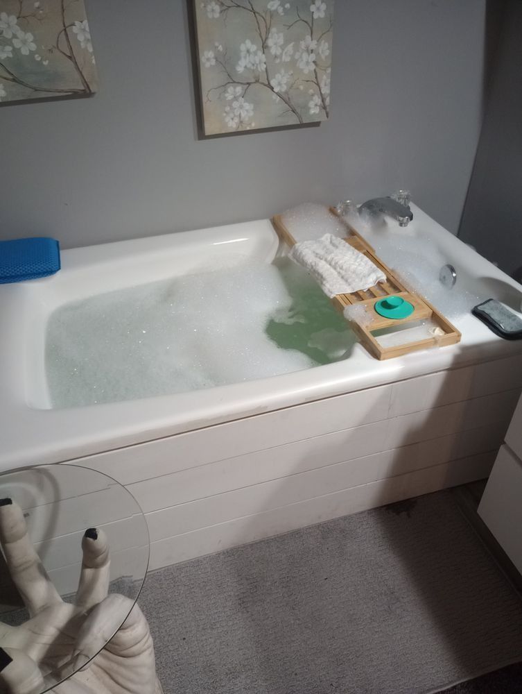 A bubble bath in a white tub with a wooden tray, a green soap dish, and a hand-shaped glass side table.