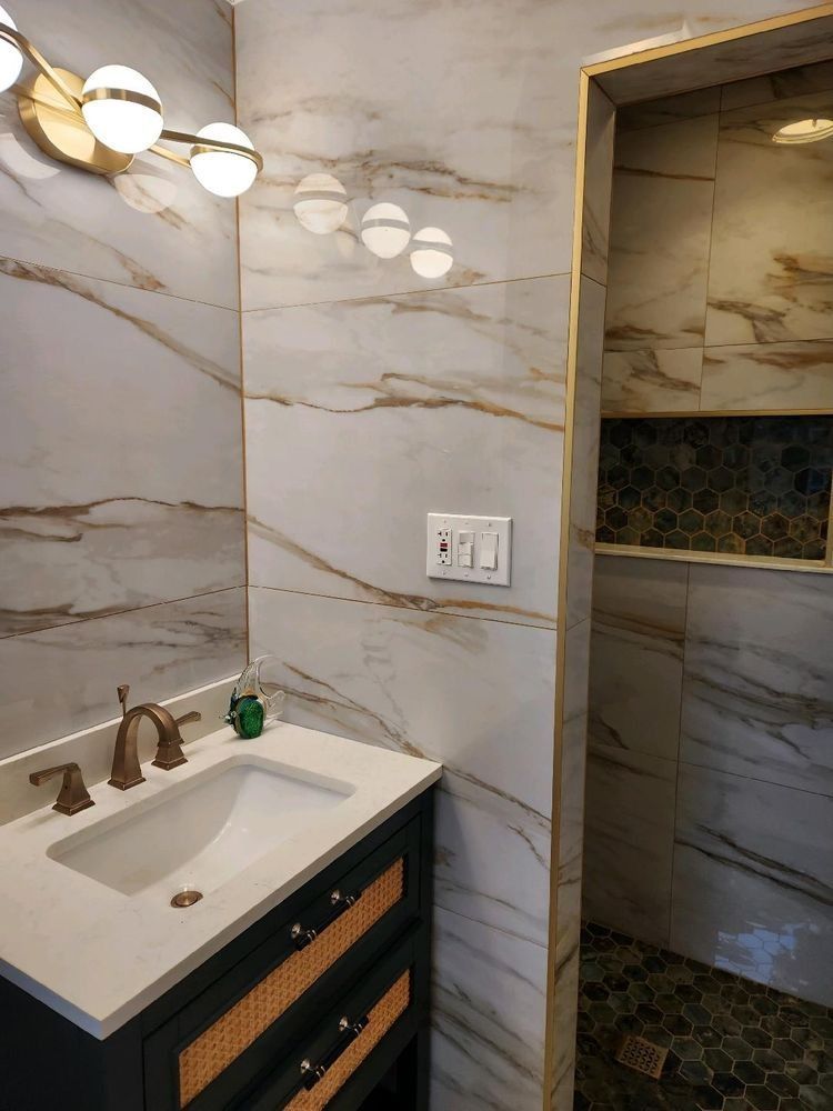 A modern bathroom vanity with a white sink, gold faucet, and textured drawers against marbled walls, next to a shower area.