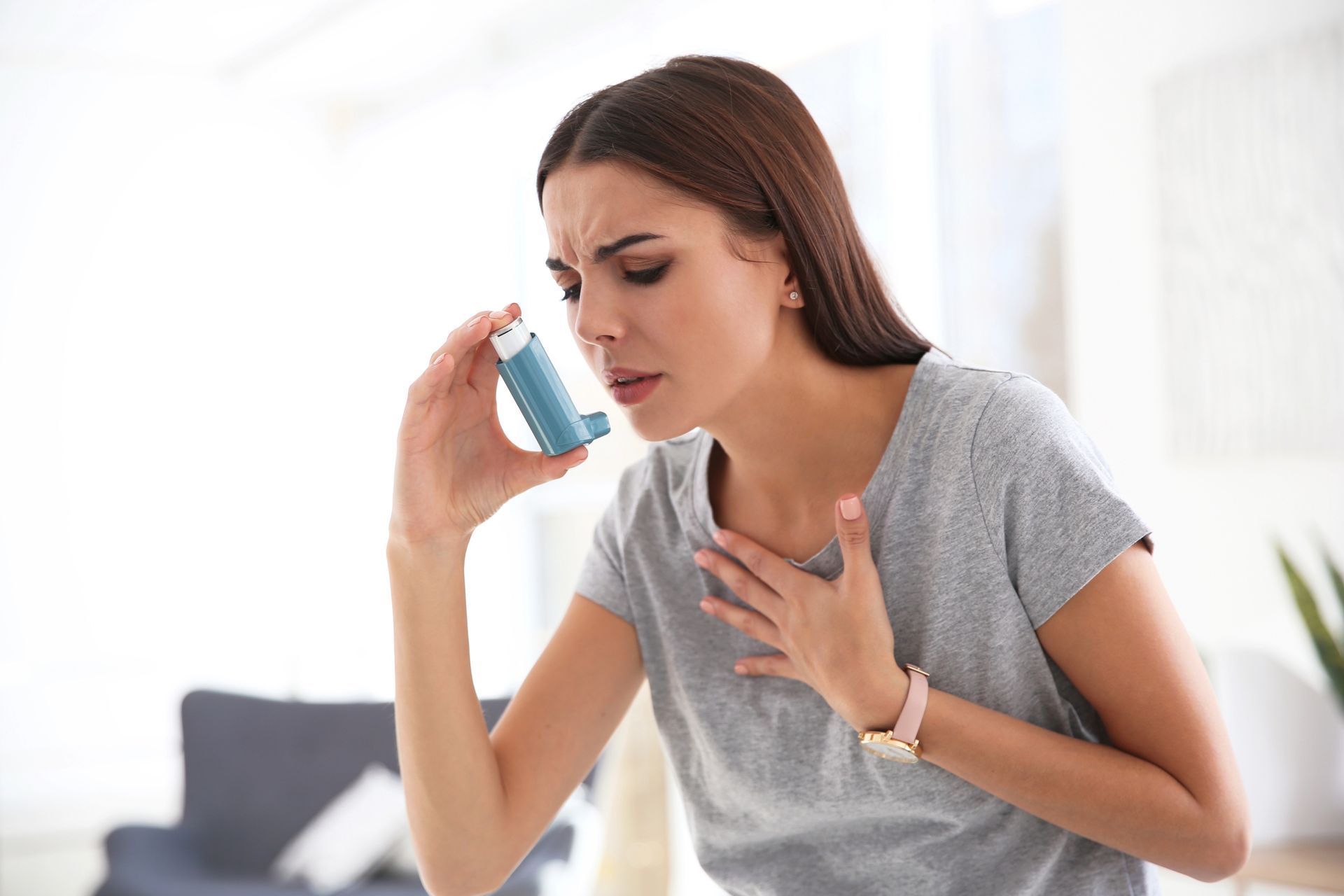 Woman holding inhaler, looking distressed, hand on chest, indoors.