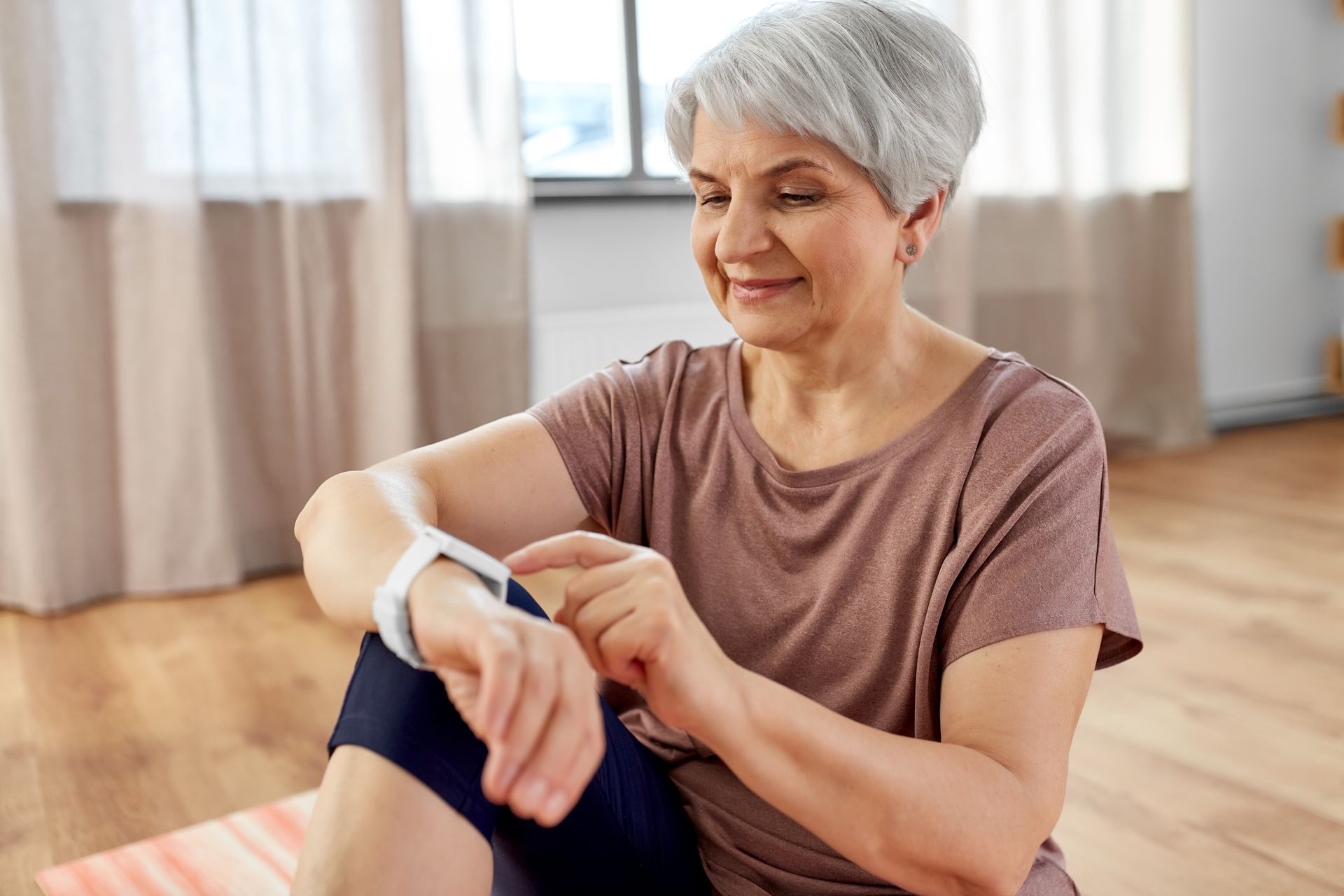 An elderly woman is sitting on a yoga mat and looking at her smart watch.