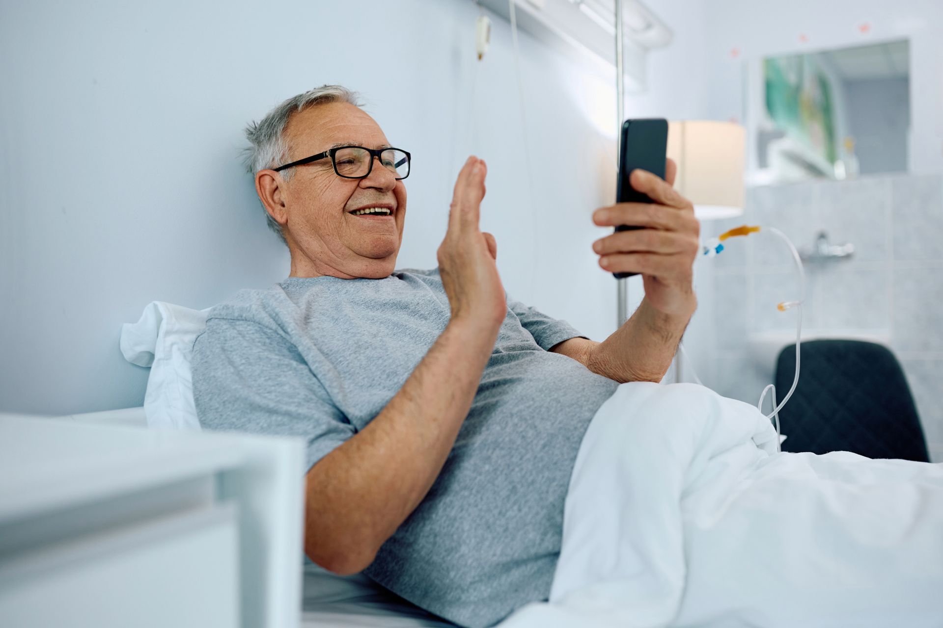 Man in hospital bed waves at phone during video call.