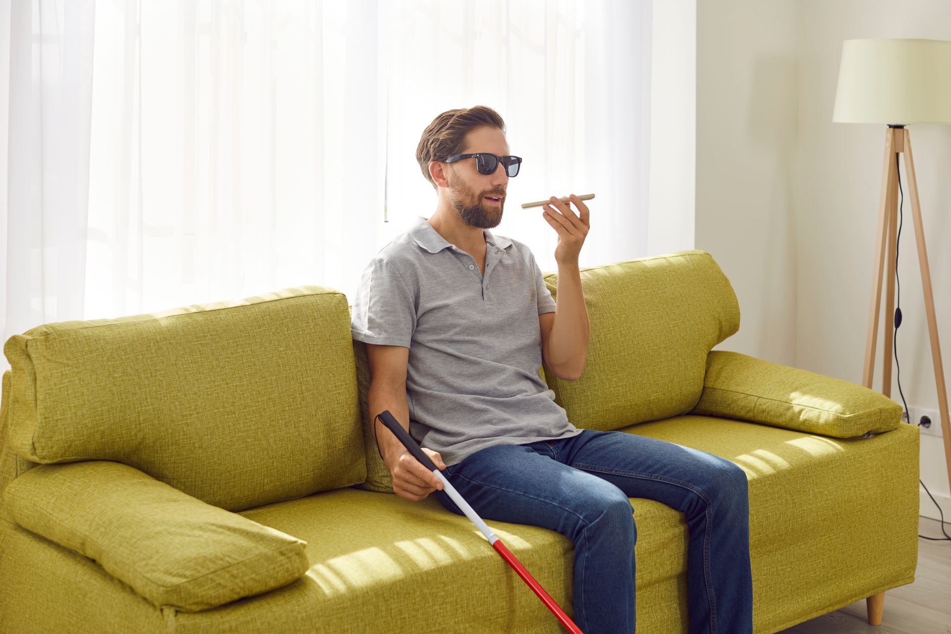 A blind man is sitting on a couch with a cane and talking on a cell phone.