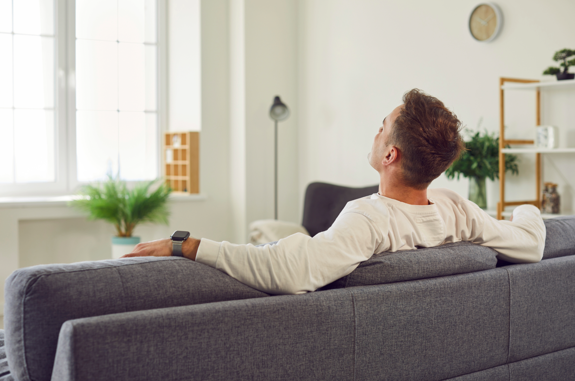 Man relaxing on a gray couch, arms outstretched, looking upward in a bright living room.