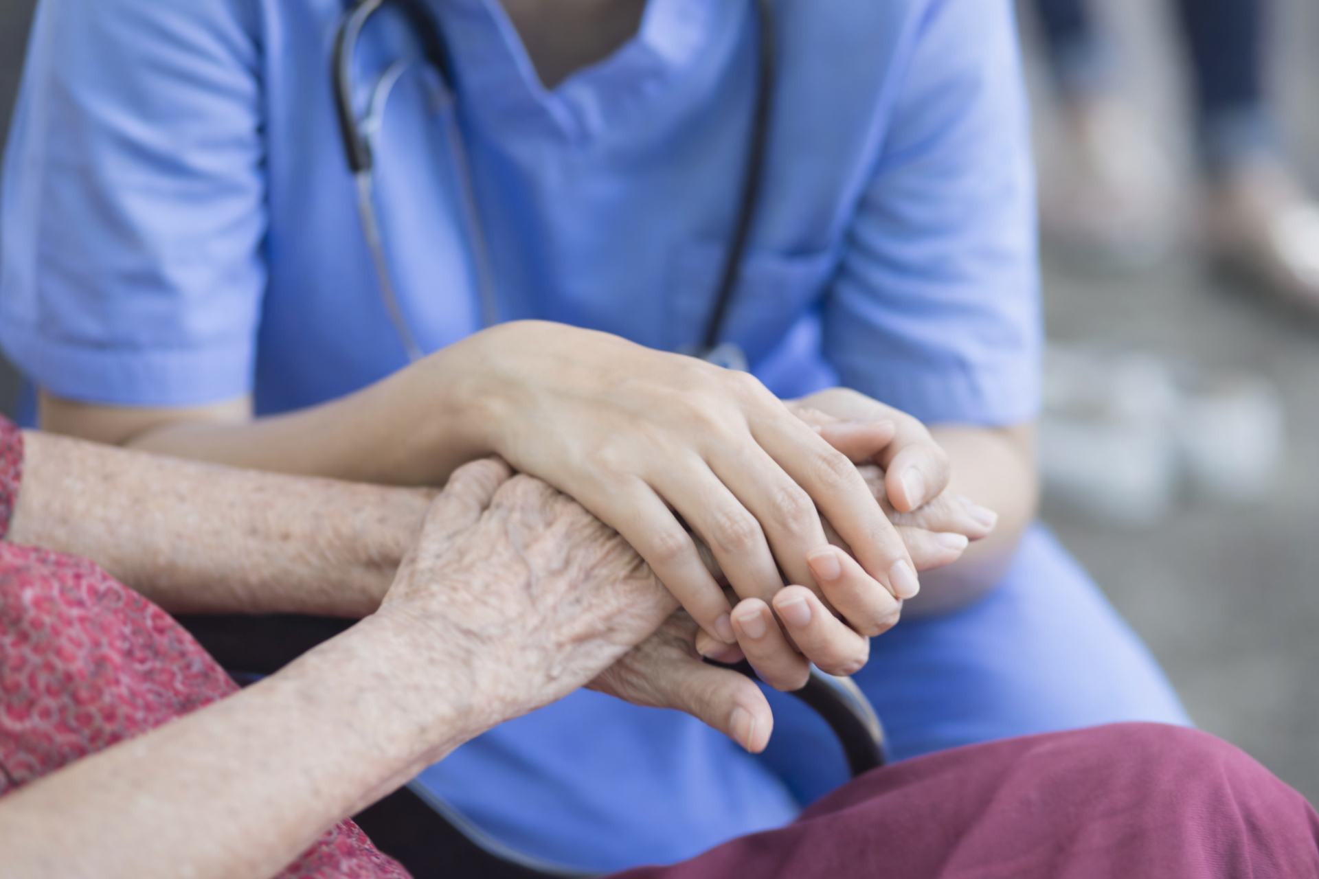 A healthcare worker in blue scrubs holding the hands of a person wearing a maroon garment to provide comfort.