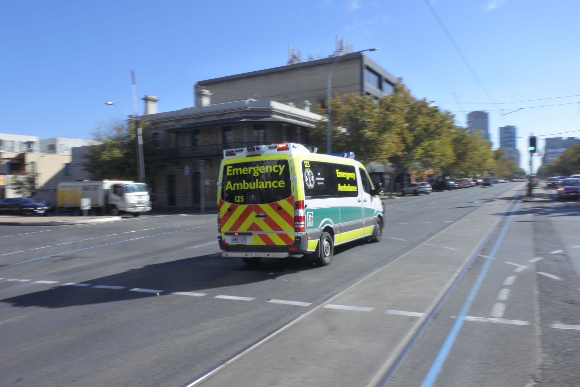 Ambulance driving on a city street. Yellow and green with emergency lights. Buildings in the background.