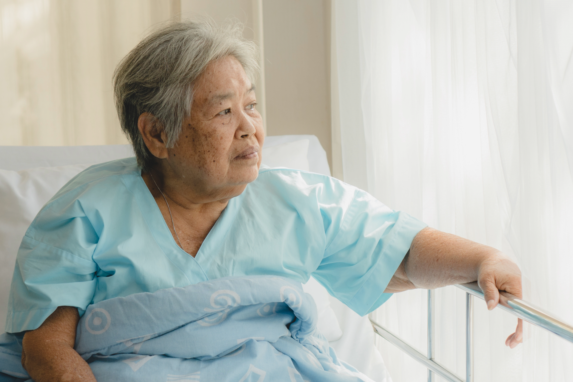 Elderly person in hospital bed, looking out window. Wearing blue hospital gown, hand on bed rail.