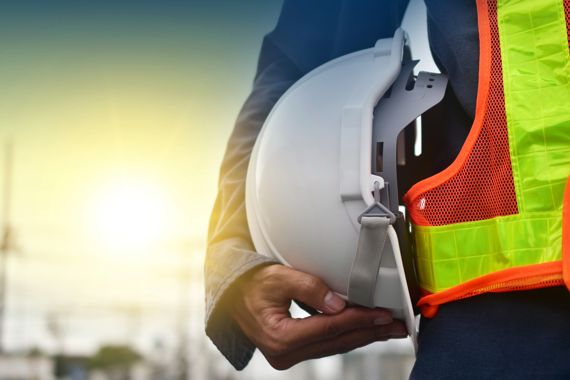 Construction worker holding a white hard hat beside a high-visibility orange and green vest at sunset