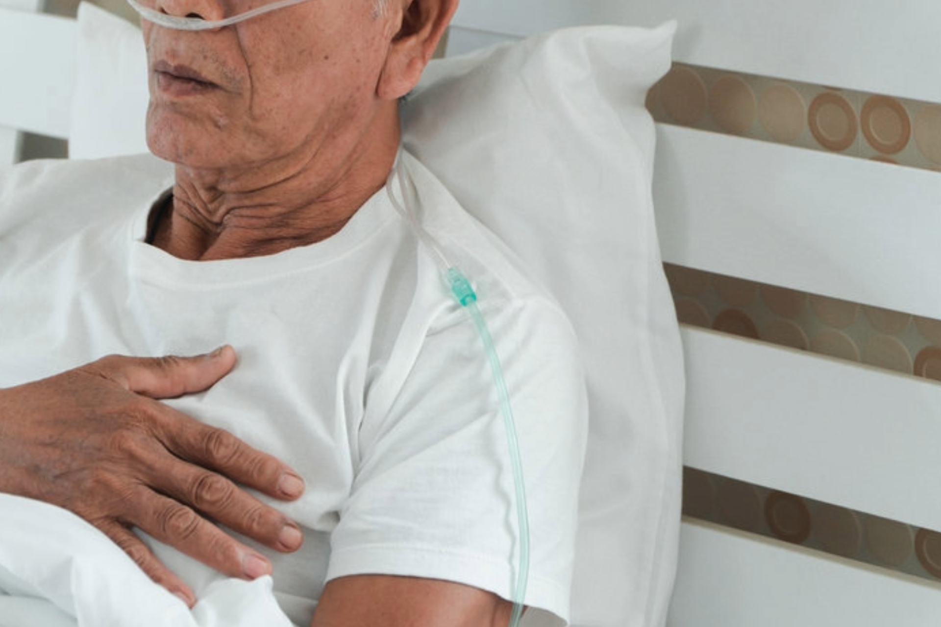 Man in hospital bed wearing oxygen cannula, hand on chest, resting against white pillow and striped headboard
