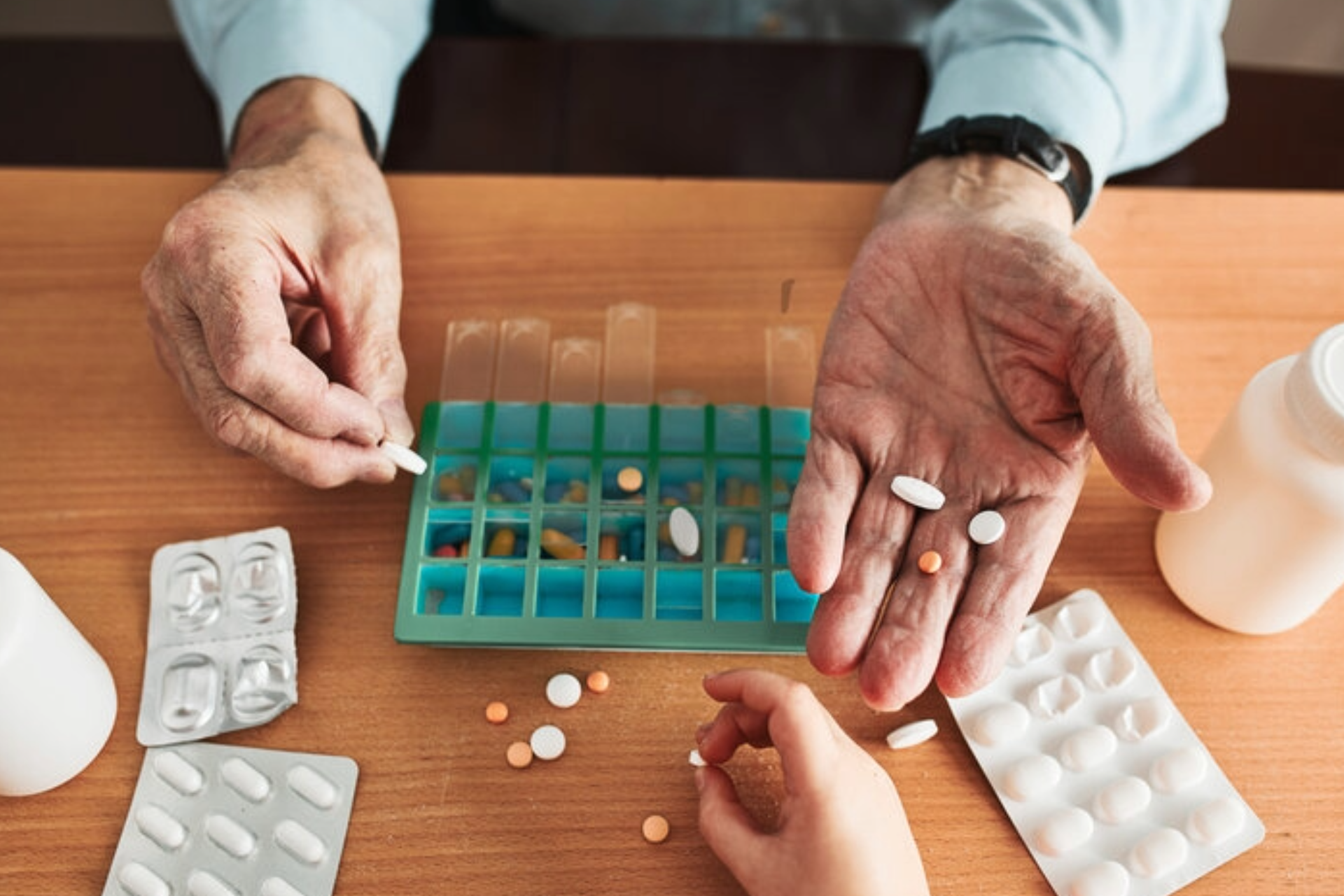 Hands sorting assorted pills into a weekly pill organizer on a wooden table.