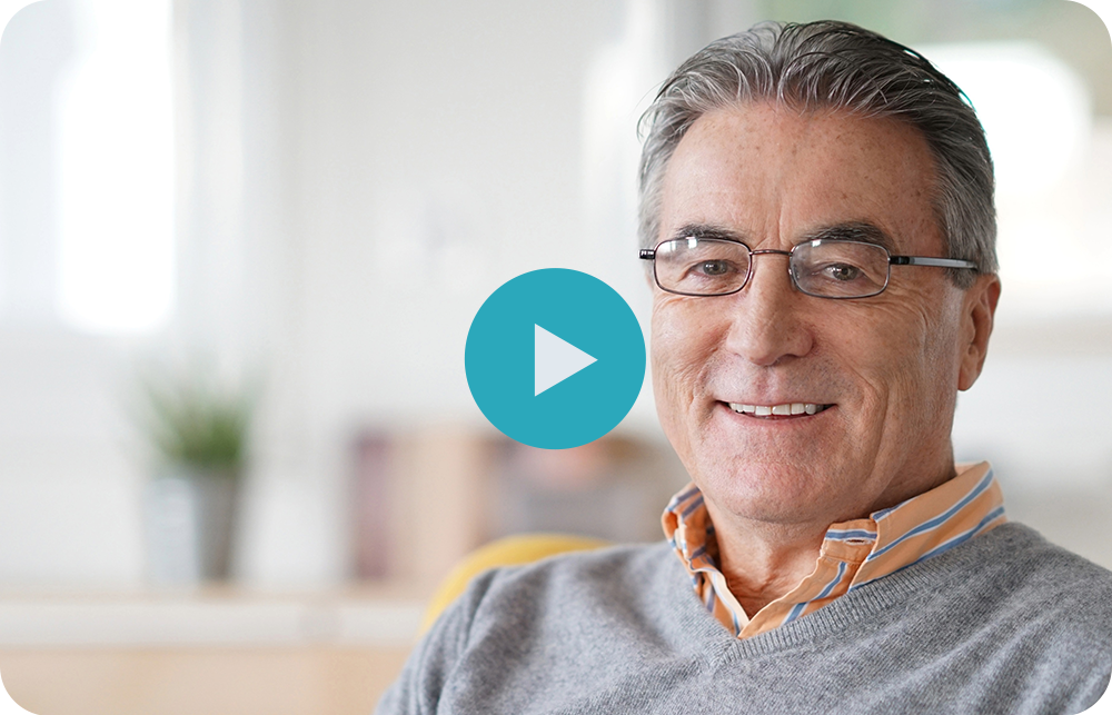 Smiling man with glasses, wearing a gray sweater and orange-striped shirt. A bright living space is in the background.