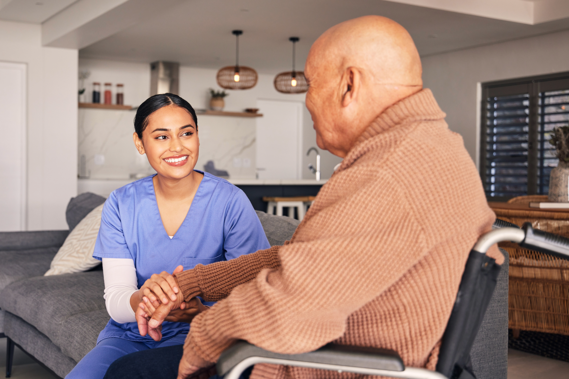 Caregiver smiling and holding hands with a seated man in a living room kitchen area