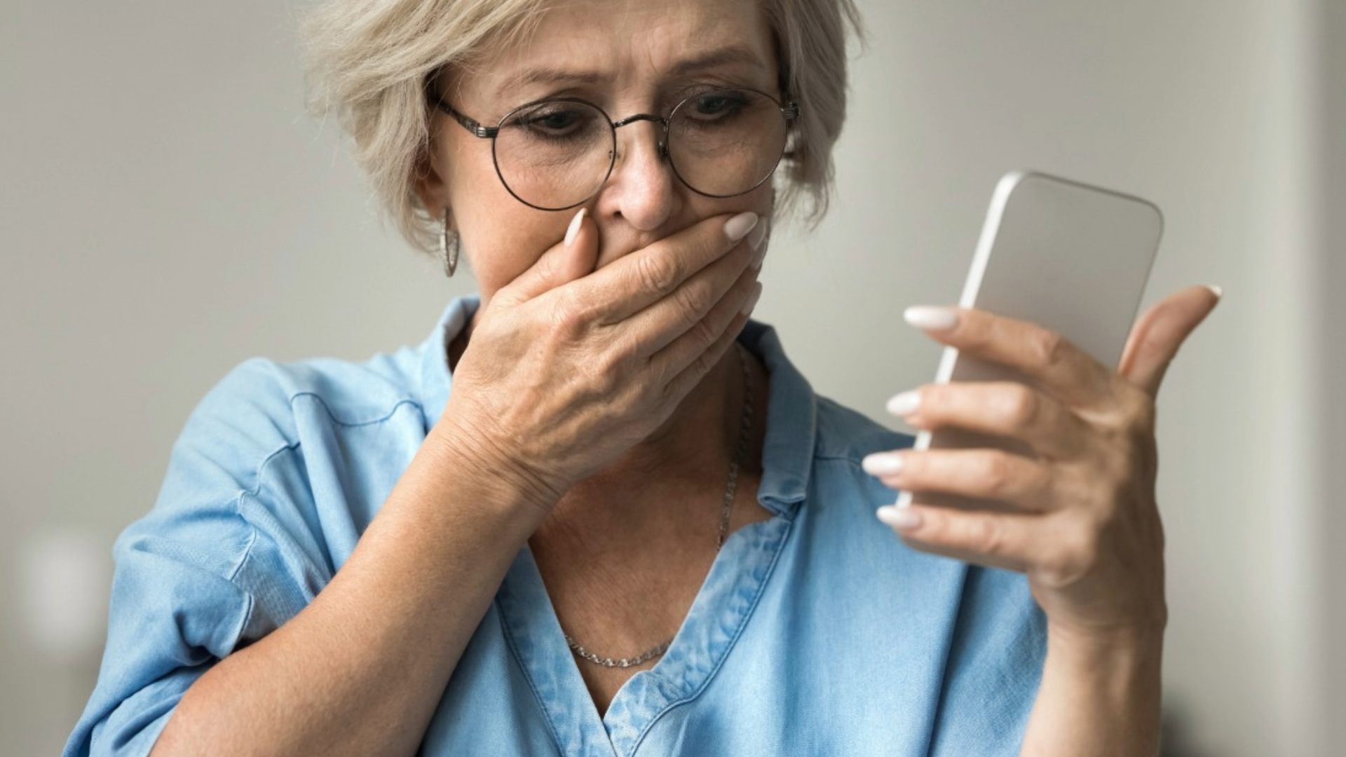 Person in glasses covering their mouth while looking at a smartphone, appearing shocked.