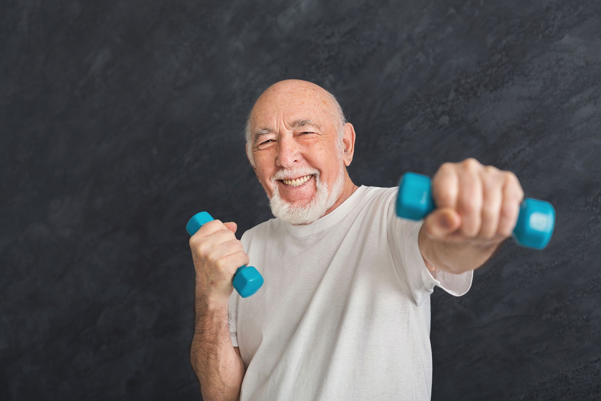 An elderly man is holding two blue dumbbells in his hands and smiling.