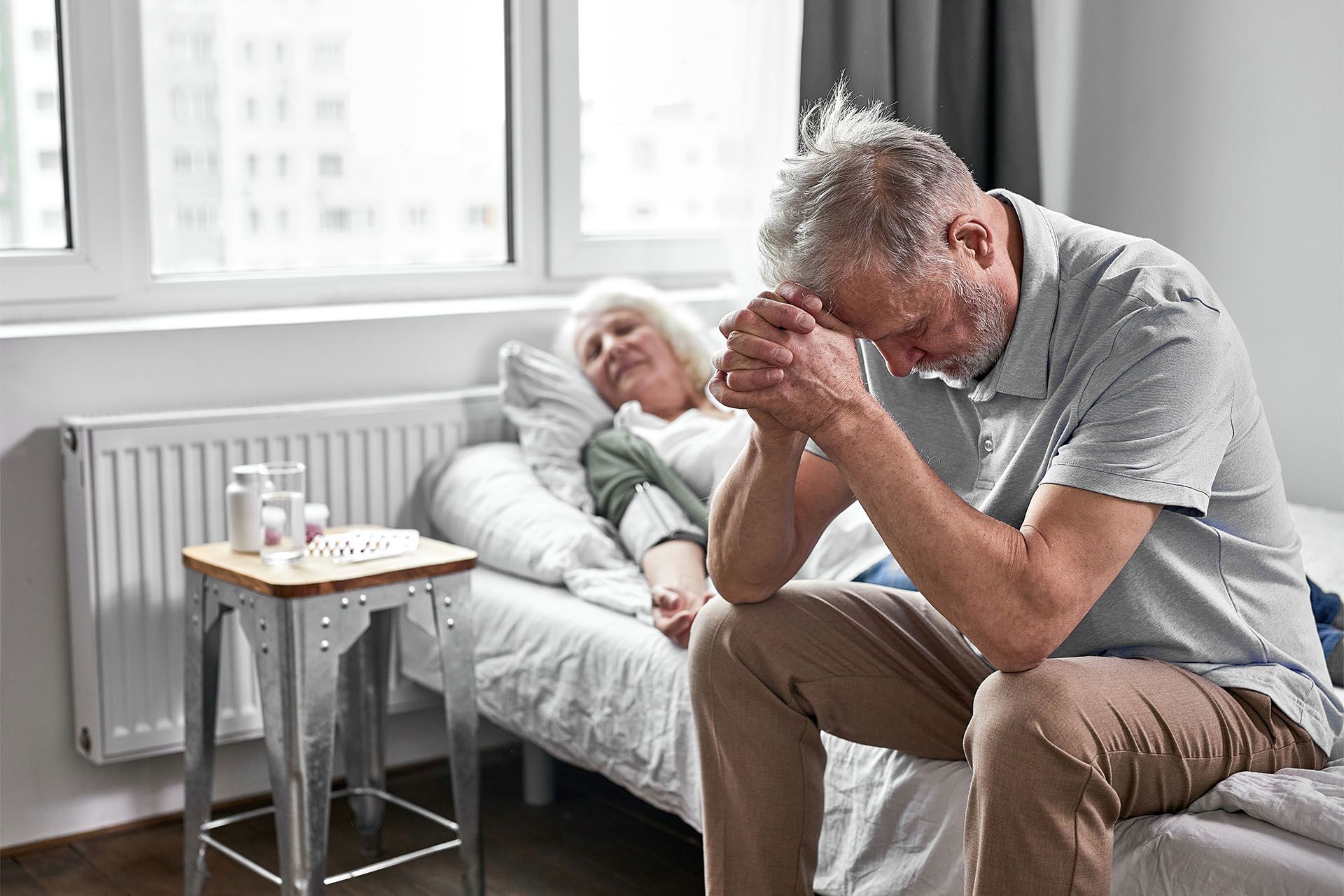 An elderly man is sitting on a bed next to an elderly woman in a hospital bed.