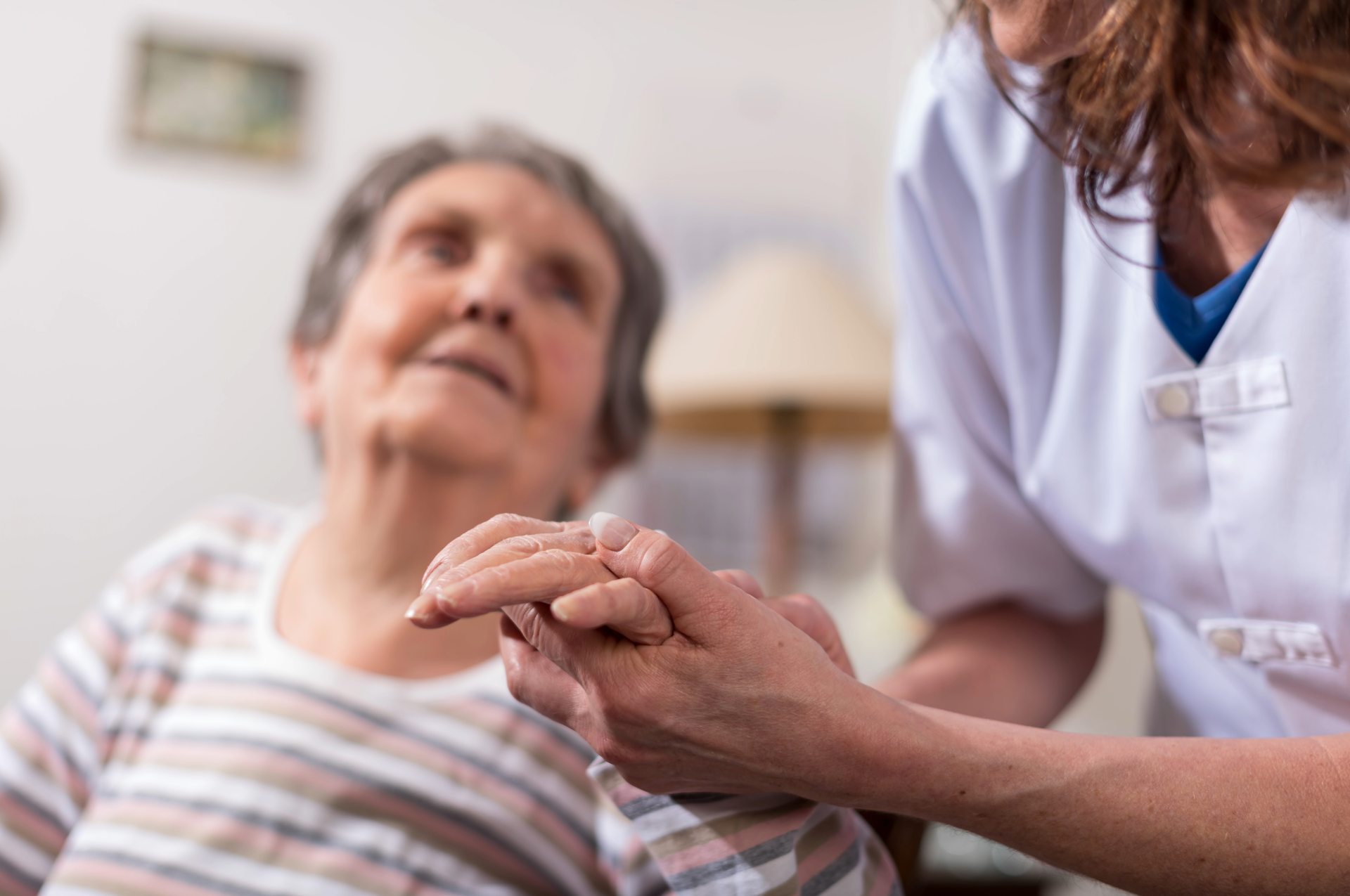 A caregiver holding hands with an elderly woman, inside a home, providing support and comfort.