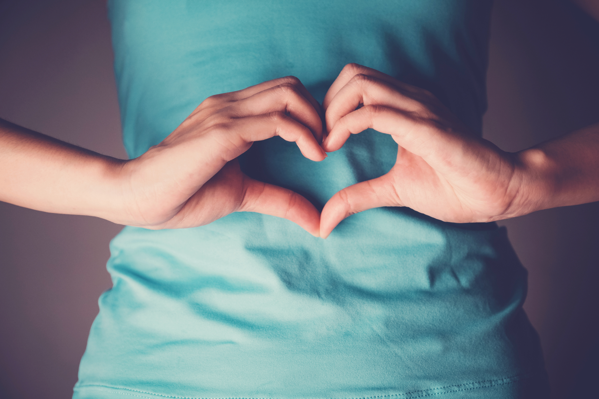 Hands forming a heart over a teal shirt on a pink background