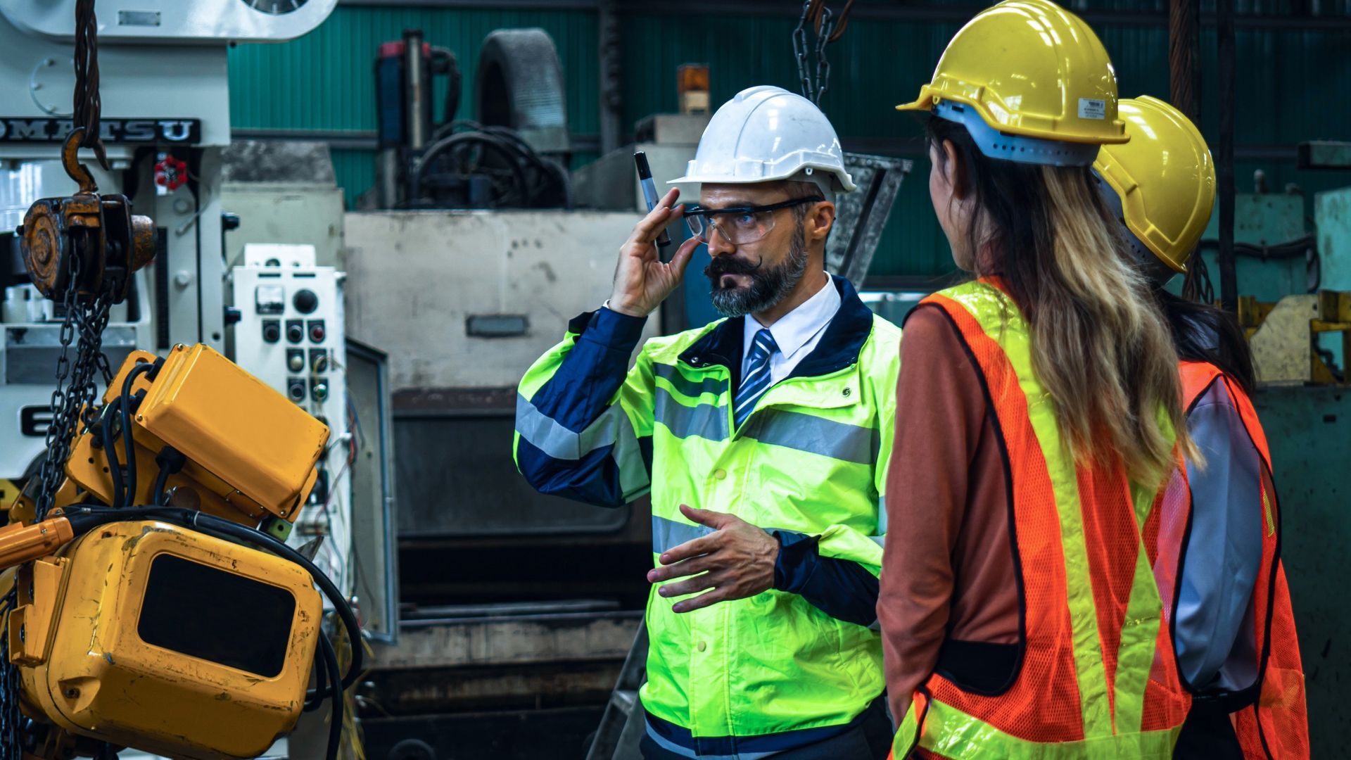 Three people in hard hats and safety vests discussing machinery in a factory.
