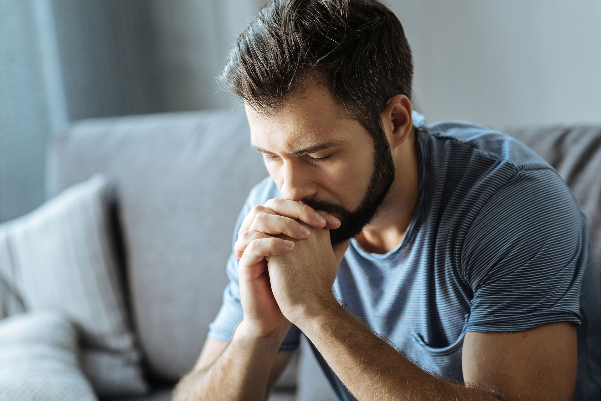 A man with a beard is sitting on a couch with his hands folded.