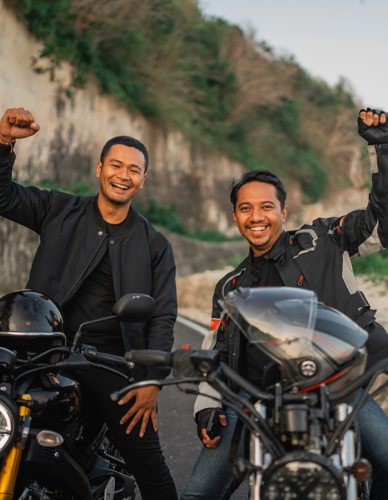 Two men on motorcycles with fists raised on a road, smiles. Cliffs in the background.