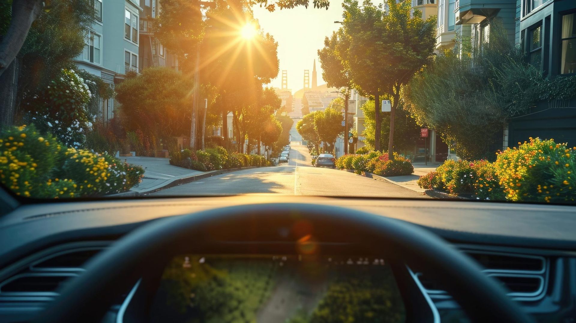 Sunlit residential street viewed from a car windshield, with trees, flowers, and houses lining the road.