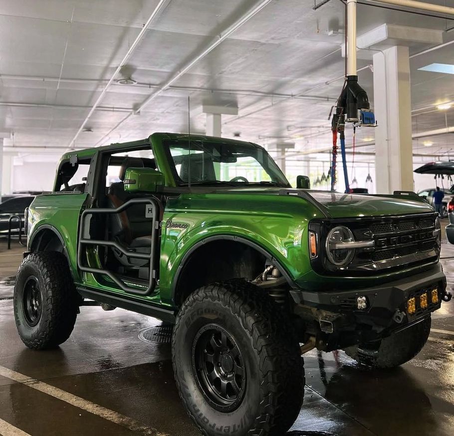 Green Ford Bronco with custom off-road modifications, including open doors with roll cages and large tires, in a garage.