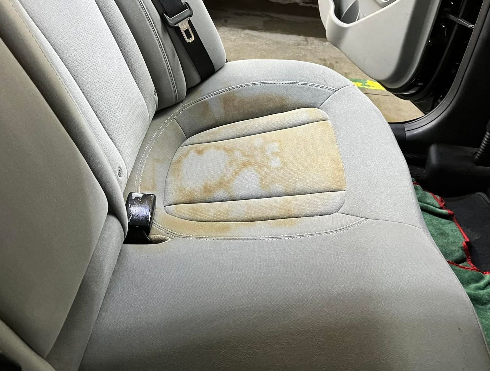 Car back seat with large, discolored stain on light gray fabric. A seatbelt is visible.