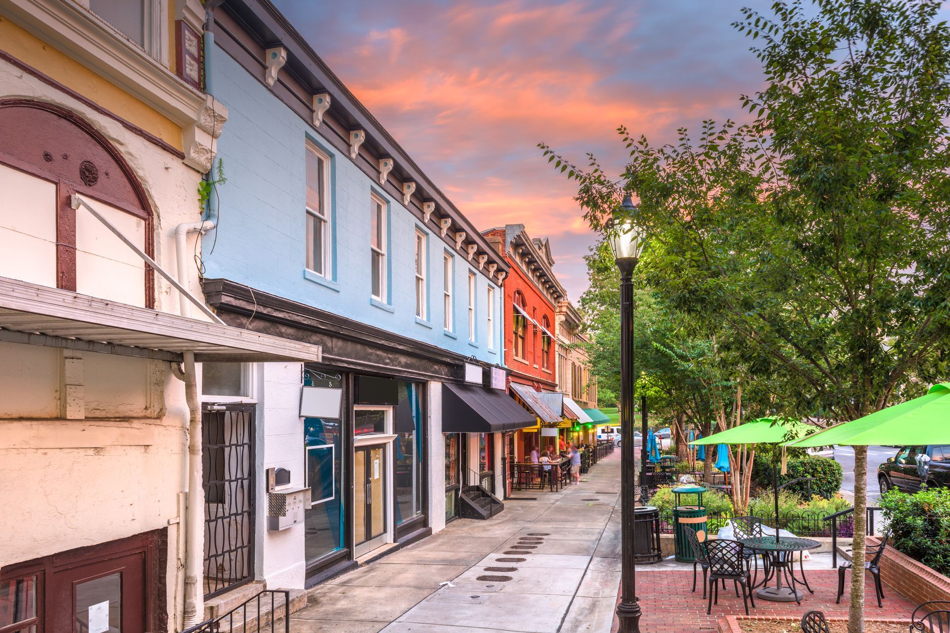 Colorful storefronts lining a quiet pedestrian street at sunset, with café tables and green umbrellas on the right.