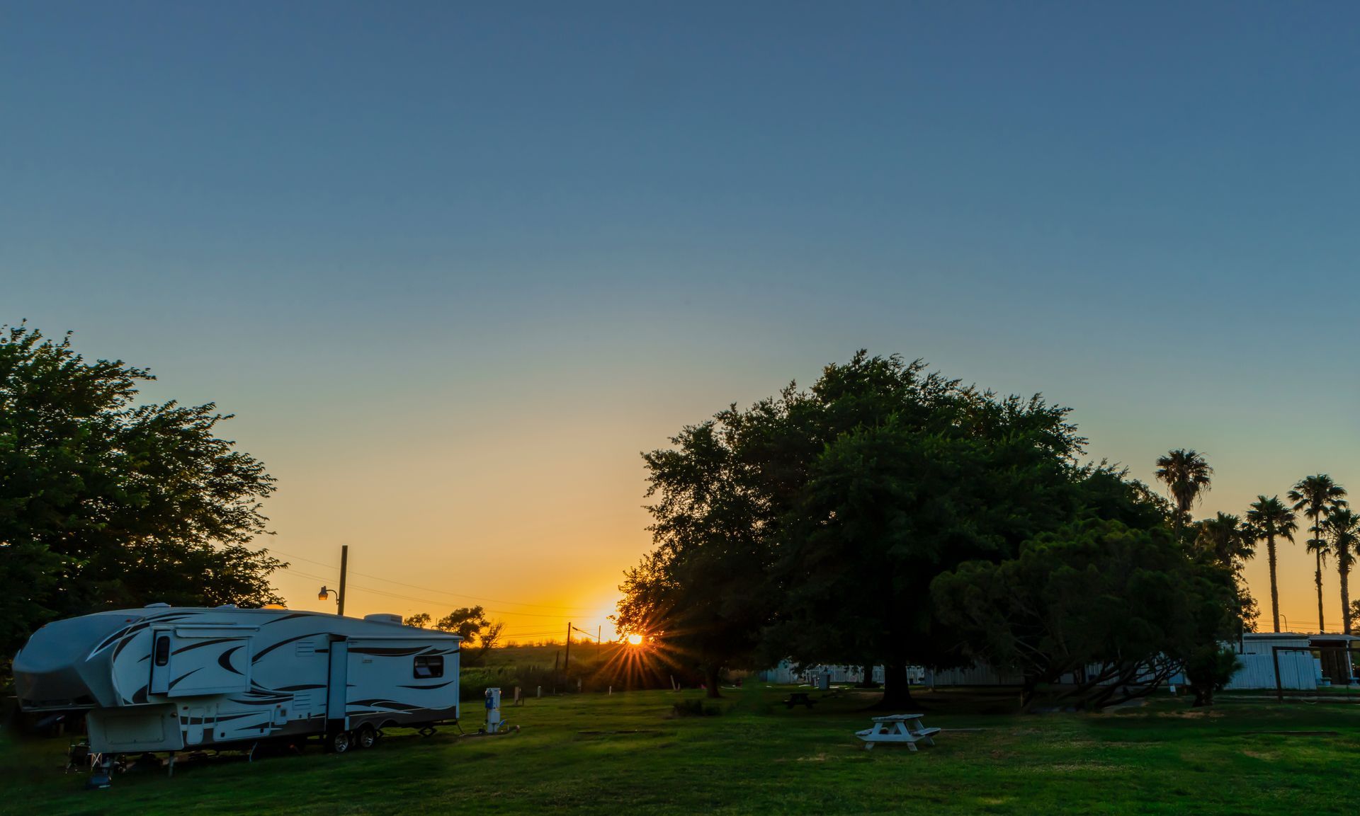 Sunset over a grassy campground with a camper trailer, trees, and a clear blue-orange sky.