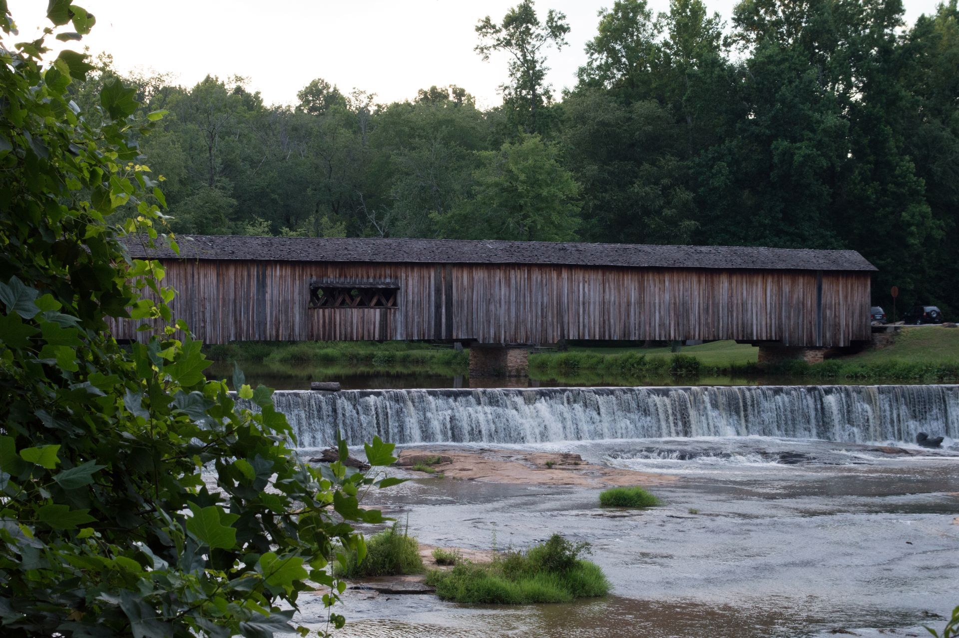 Old wooden covered bridge over a low waterfall beside a rocky river and dense green trees