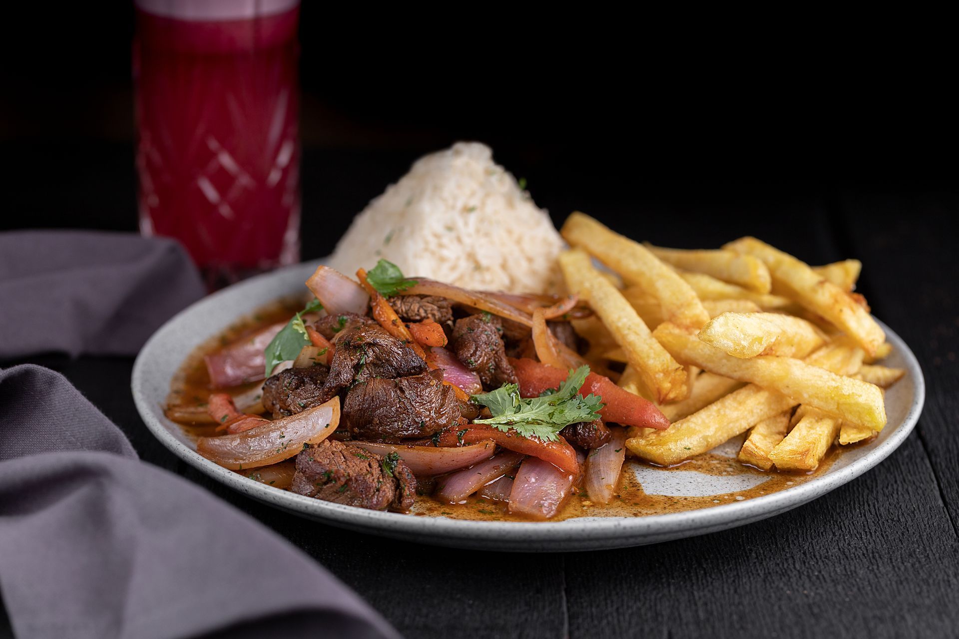 Plated grilled meat with onions, rice, and fries on a dark table, with a red drink in the background