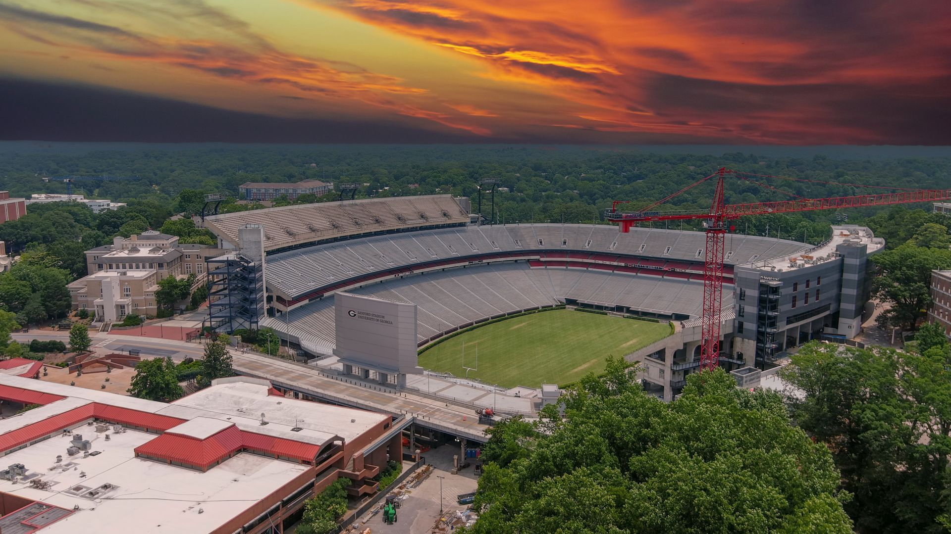 Aerial view of a stadium with a green field, surrounding buildings, and a dramatic sunset sky