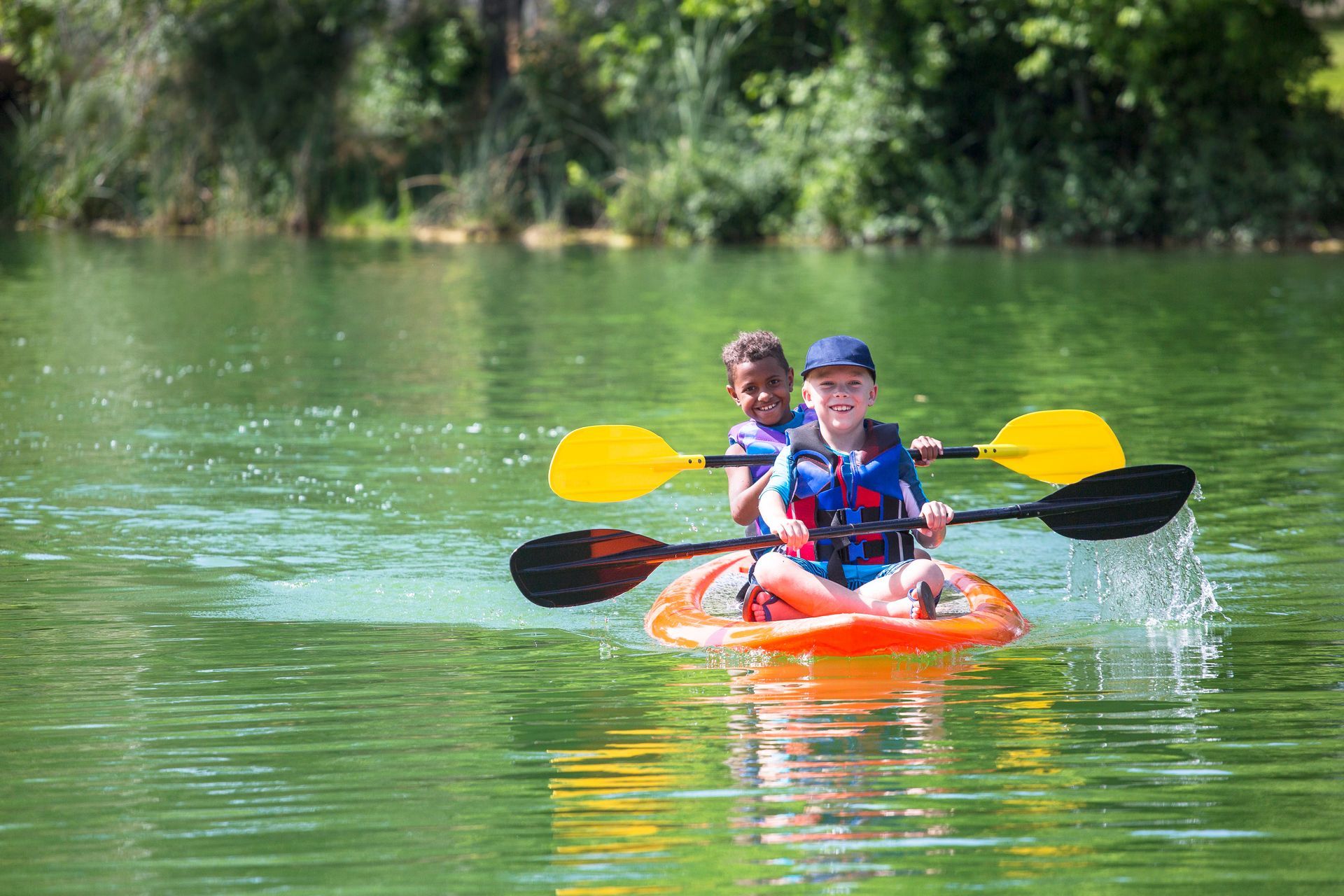 Two kayakers paddling an orange kayak on a green lake, with trees along the shore.