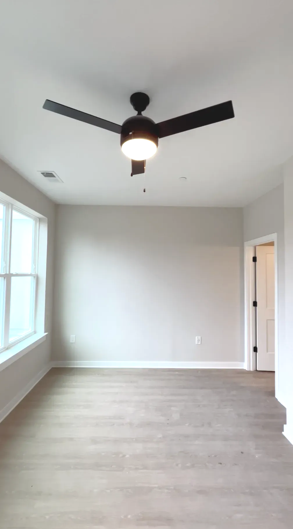 Empty apartment living room with neutral walls, light wood flooring, a ceiling fan, and large windows.