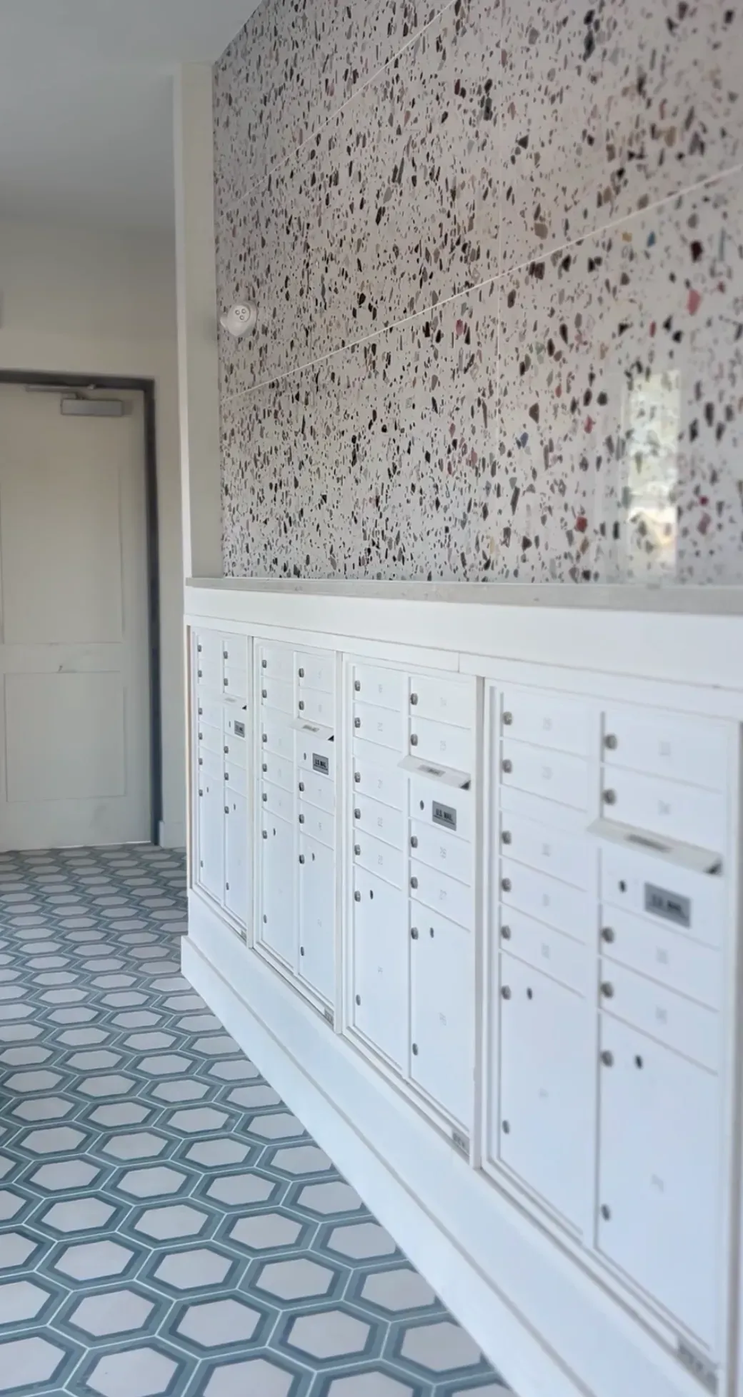 Interior building mailroom with white mailbox units along a speckled terrazzo wall.