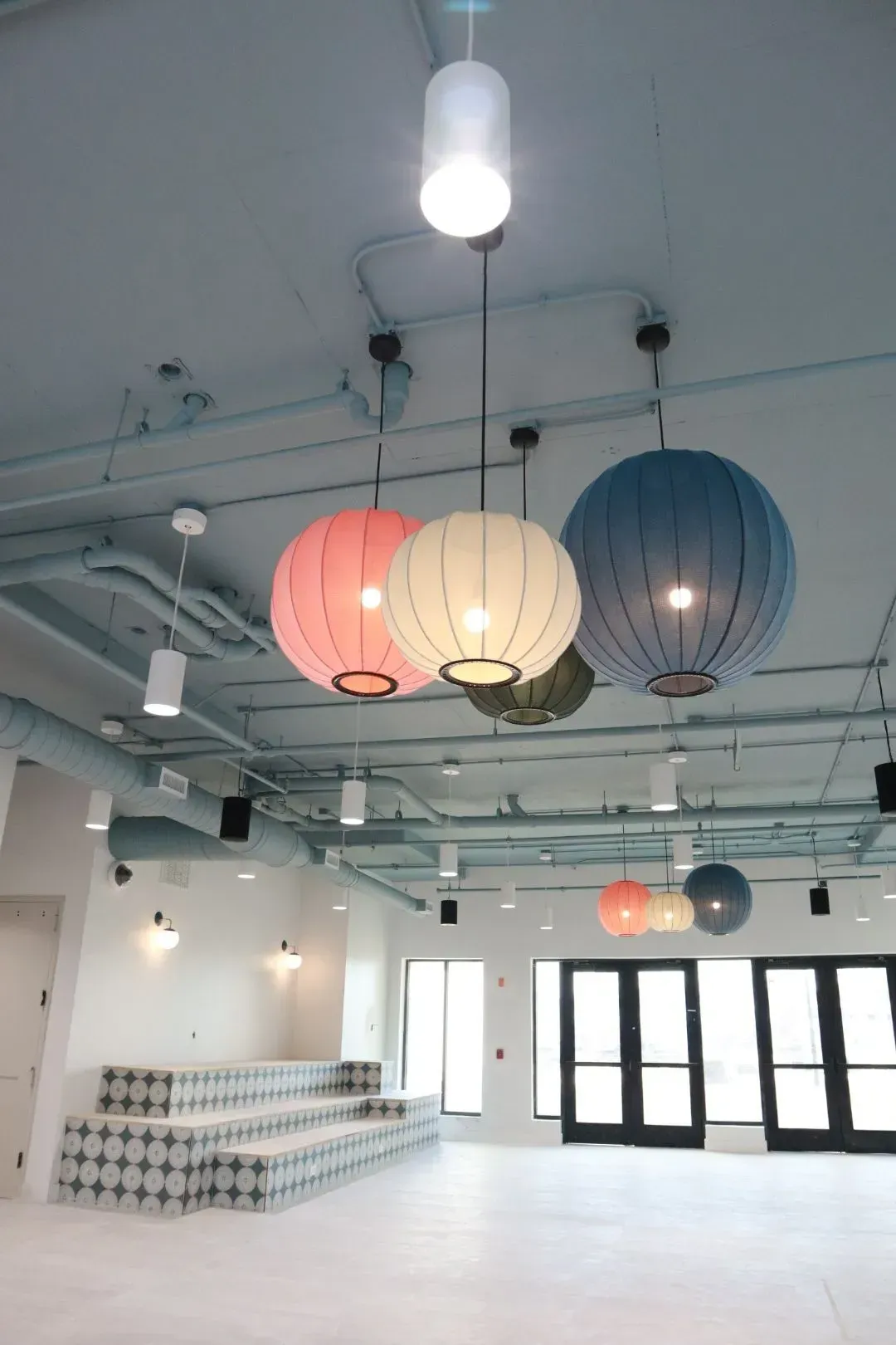 Interior community lounge with pastel hanging lanterns, built-in benches, and large glass doors.