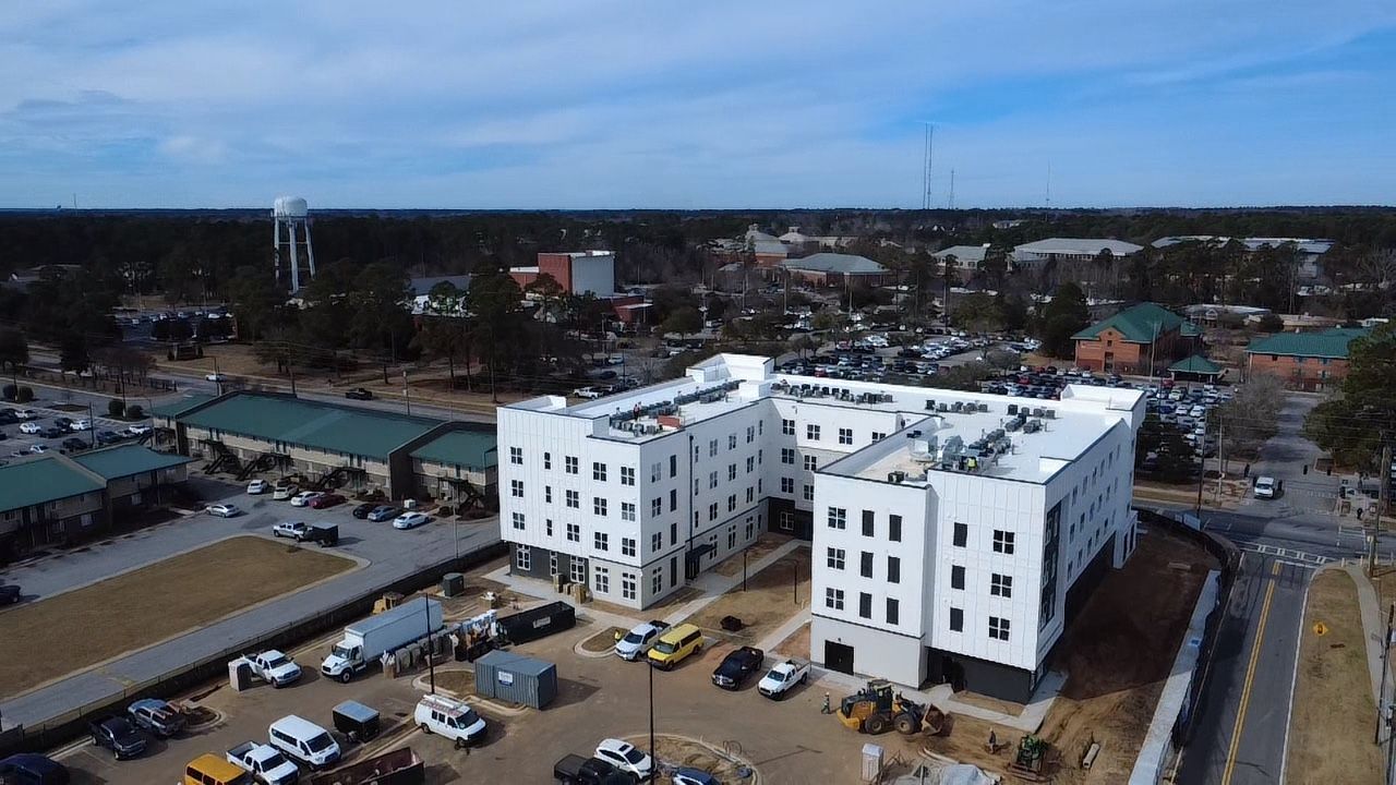 An aerial view of a white, multi-story building under construction, surrounded by parking lots and a town in the distance.