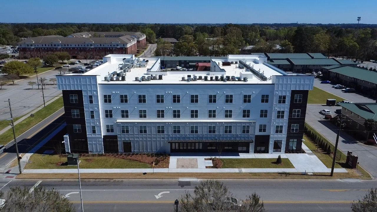An aerial view of a white, multi-story building under construction, surrounded by parking lots and a town in the distance.