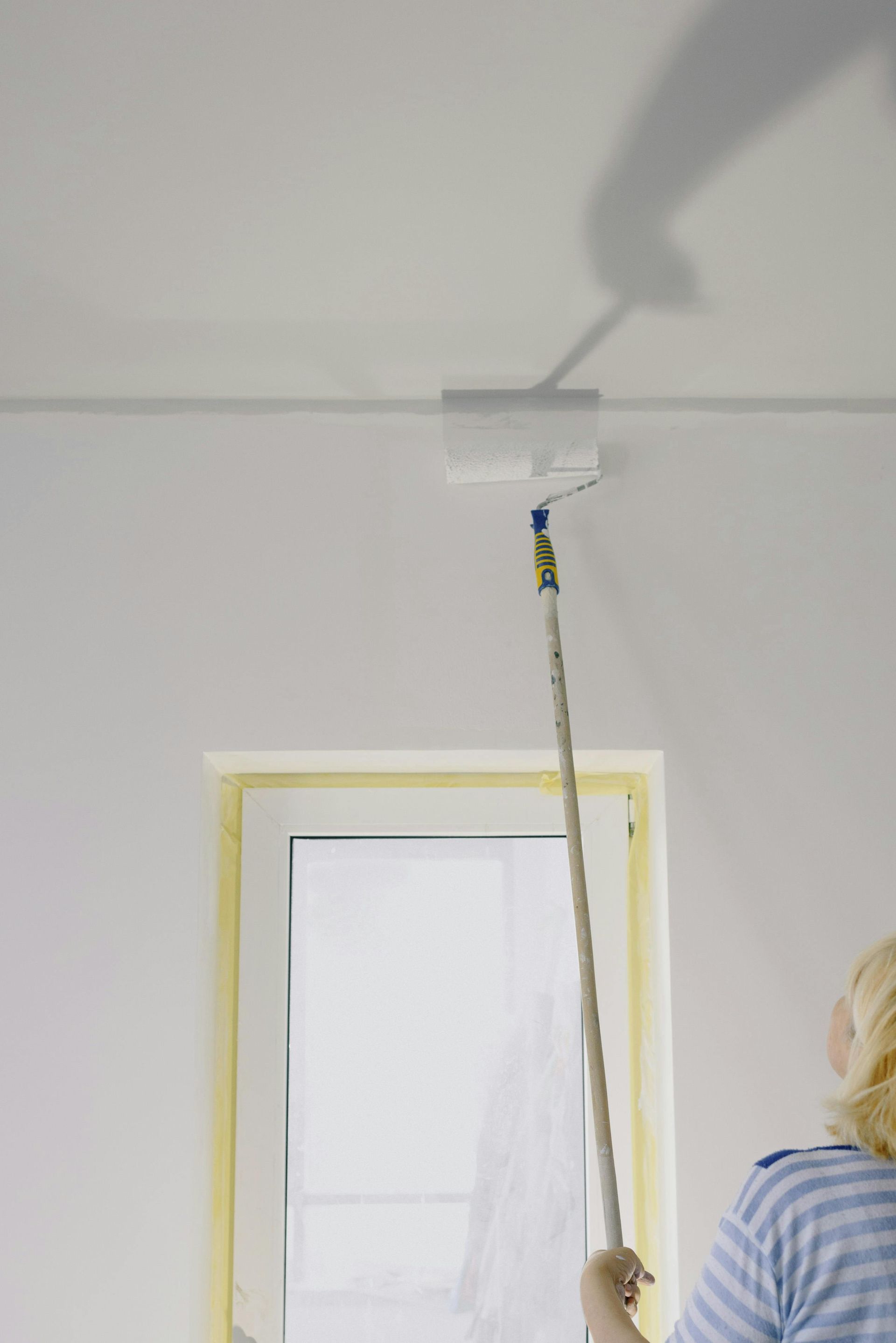 Person painting a ceiling white with a long-handled roller near a taped window opening