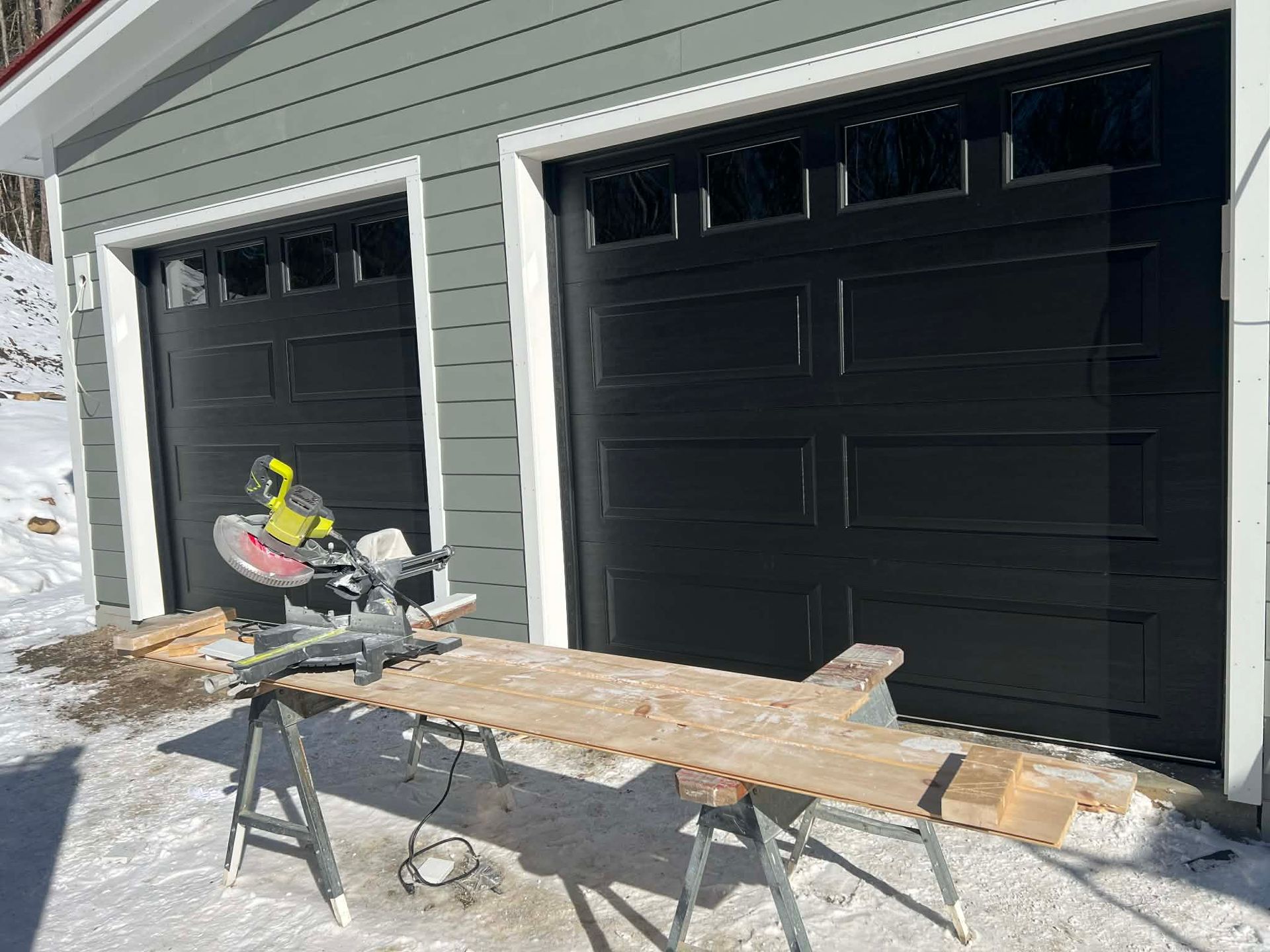 Miter saw on a workbench in front of a gray garage with black doors; snow on the ground.