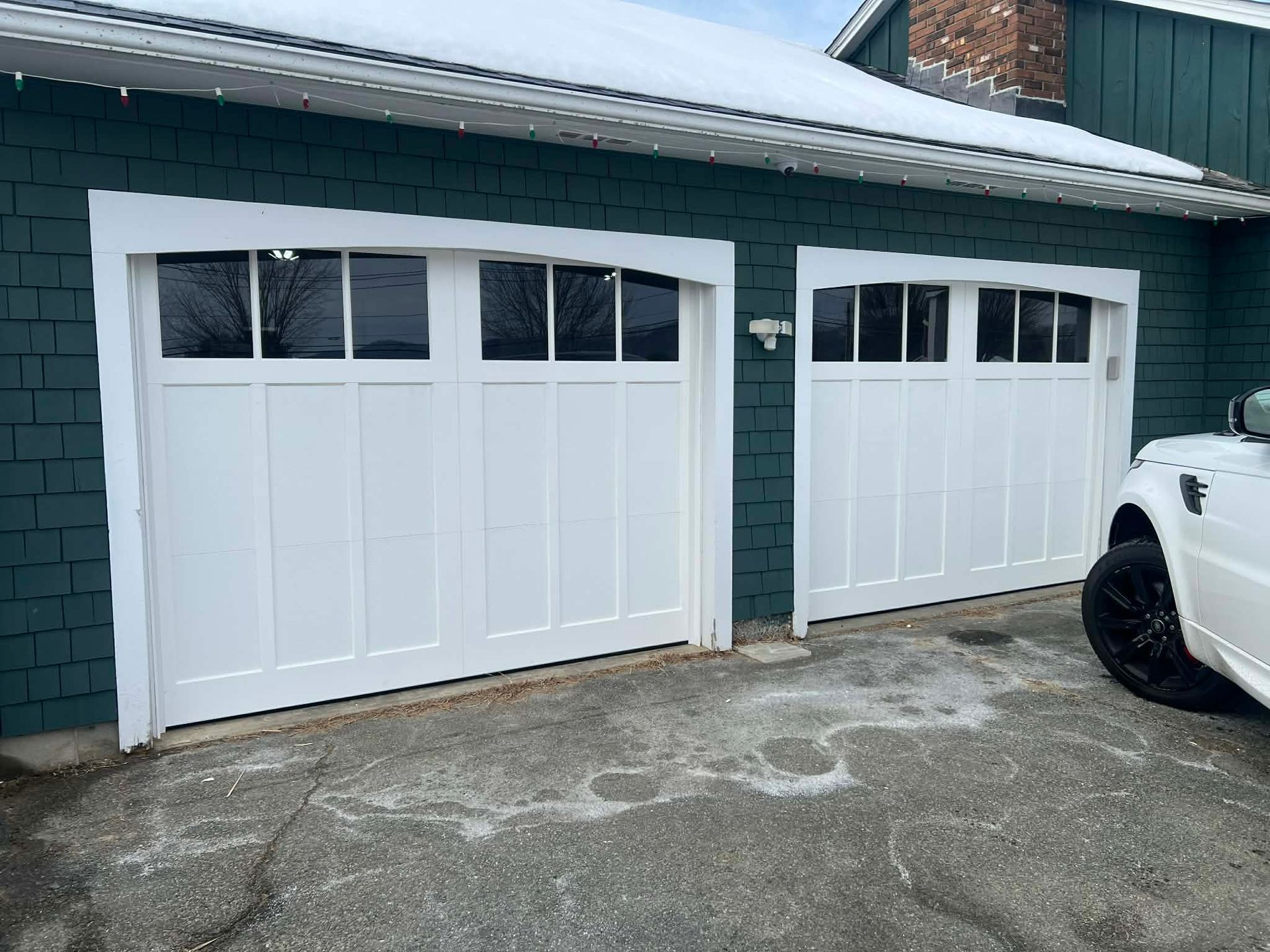 Two white garage doors with glass panels, on a teal-colored house with snow. A white vehicle is parked nearby.