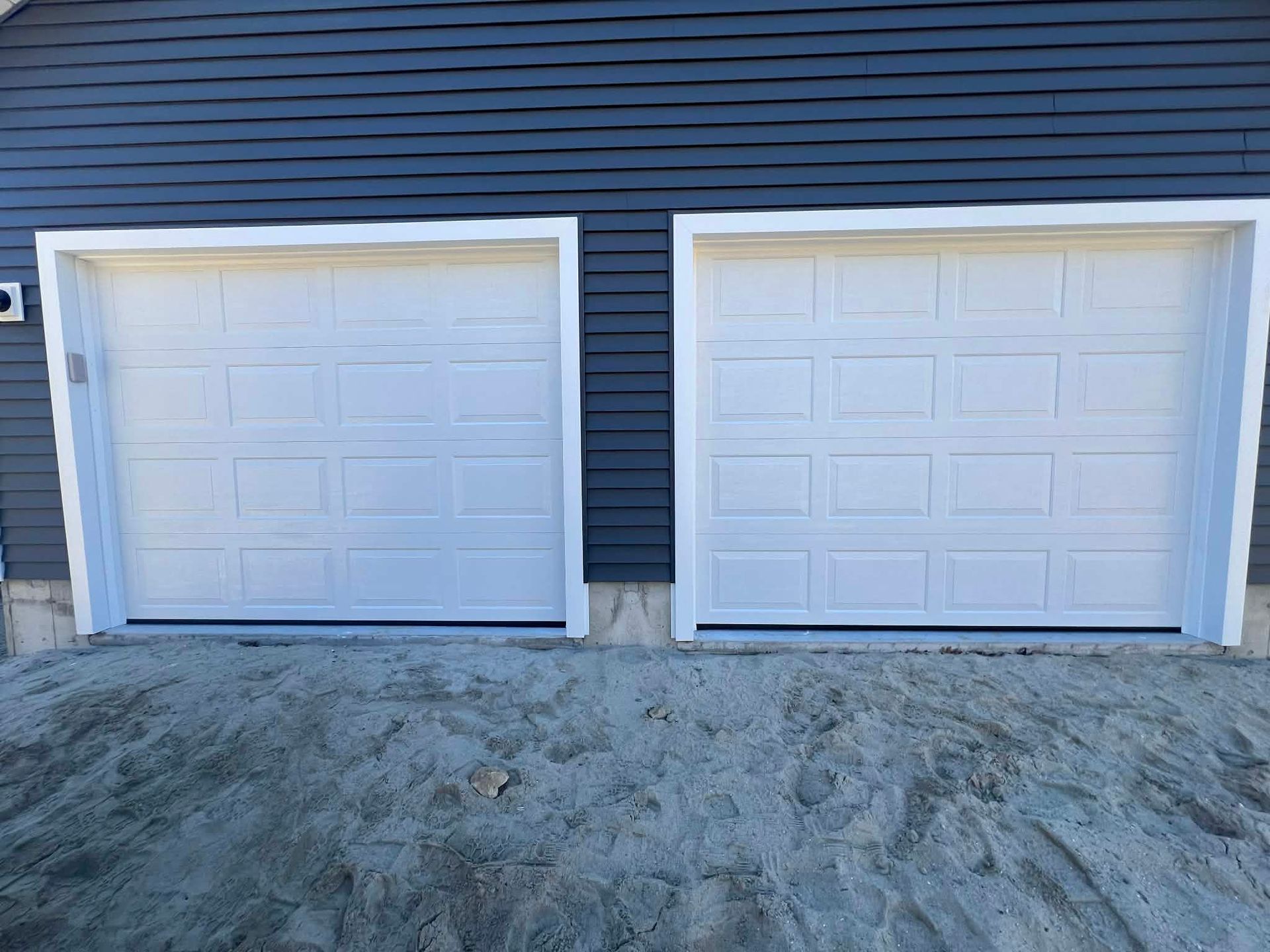 Two white garage doors with white trim, set in a blue house facade, against a gray mound.