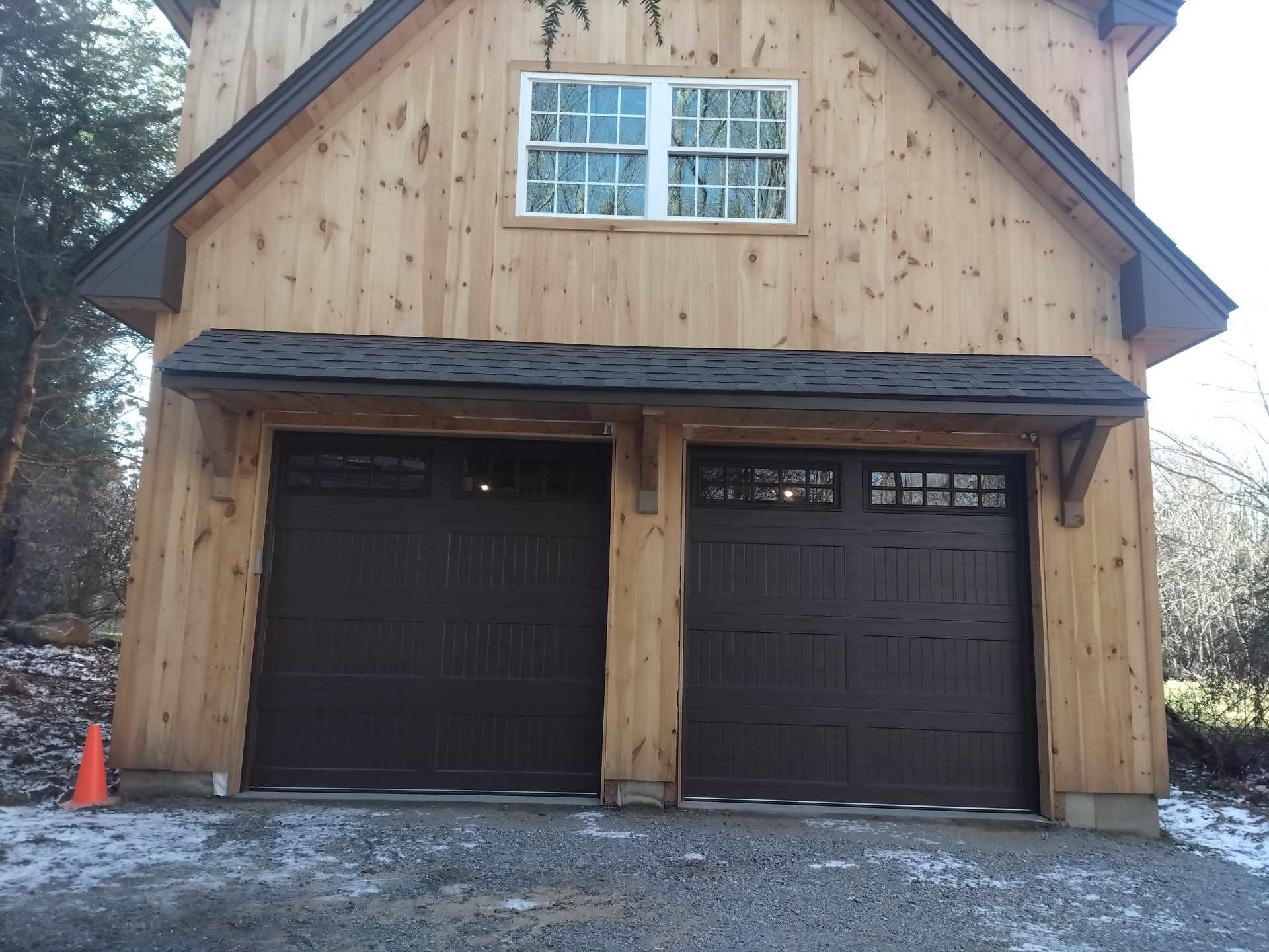 Brown two-car garage with gabled roof, window, and small overhang. Snow on the ground.