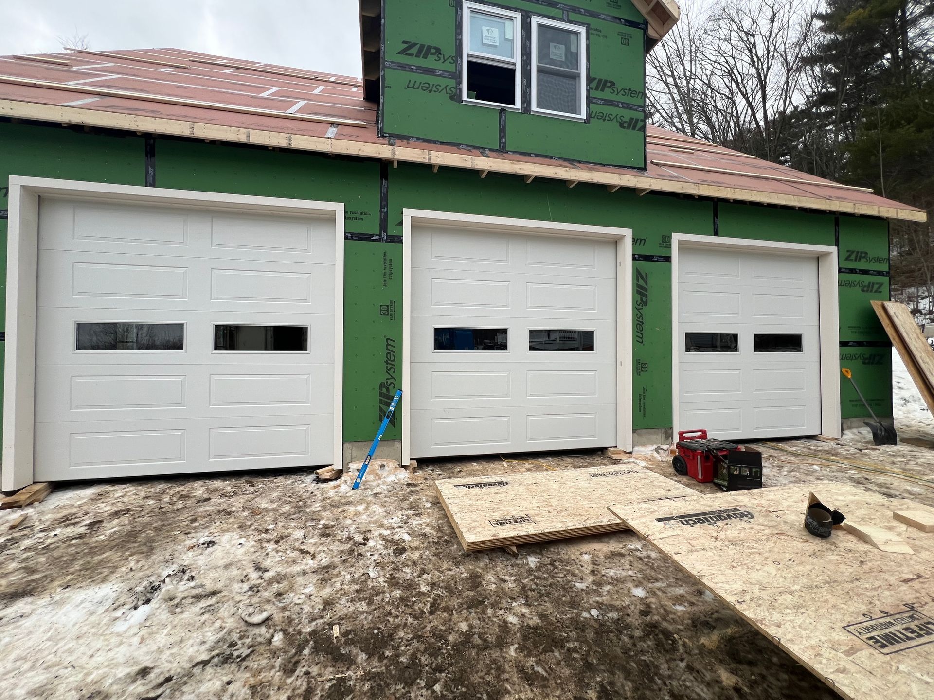 Three white garage doors with windows on a house under construction.