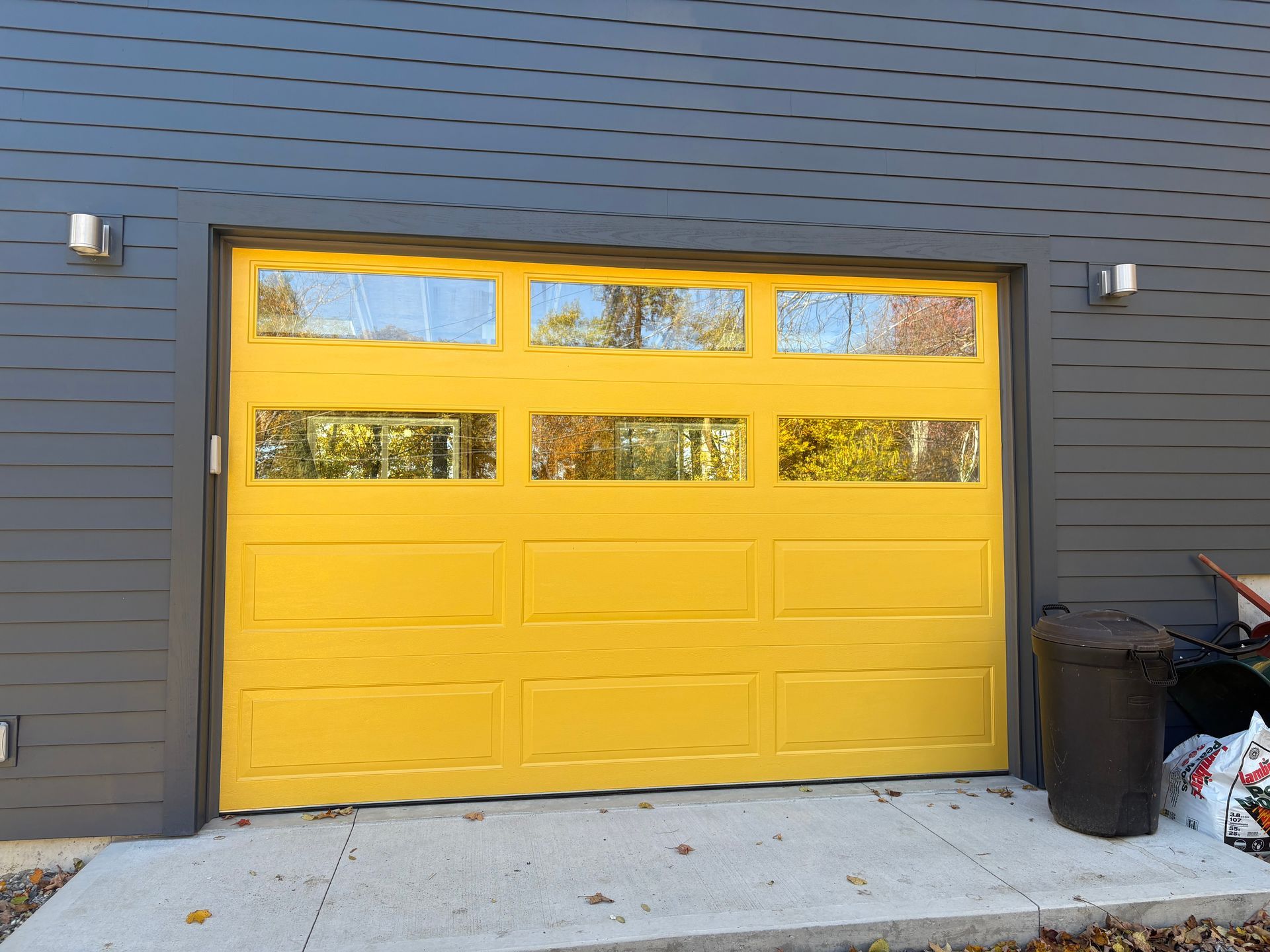 Yellow garage door with glass panes on a gray building. A trash can sits beside it.
