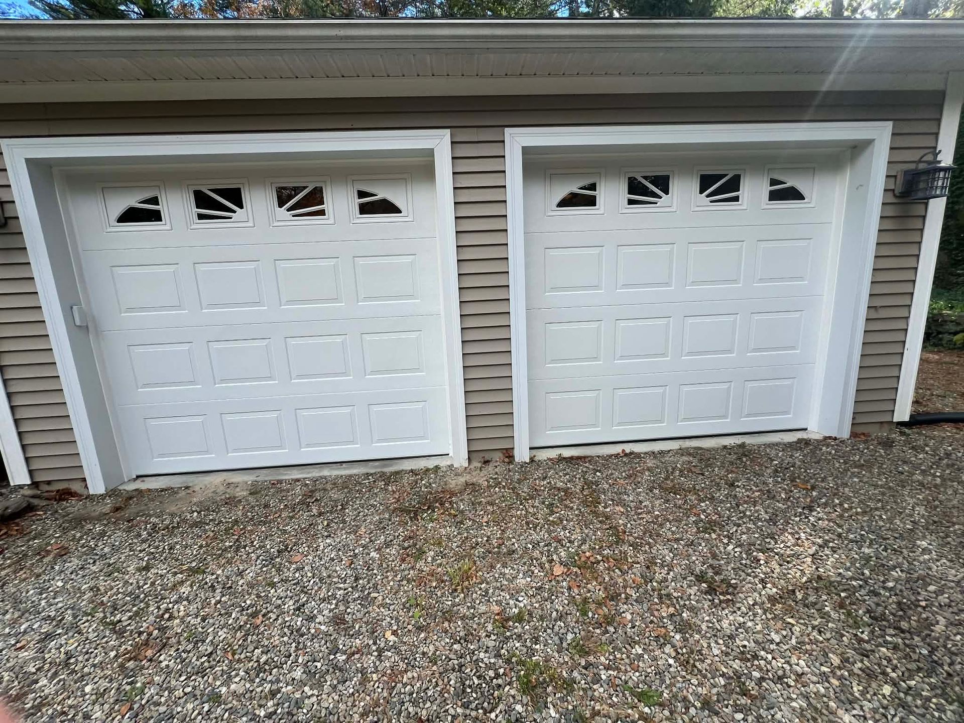 Two white garage doors with windows, set in a brown building, on a gravel surface.