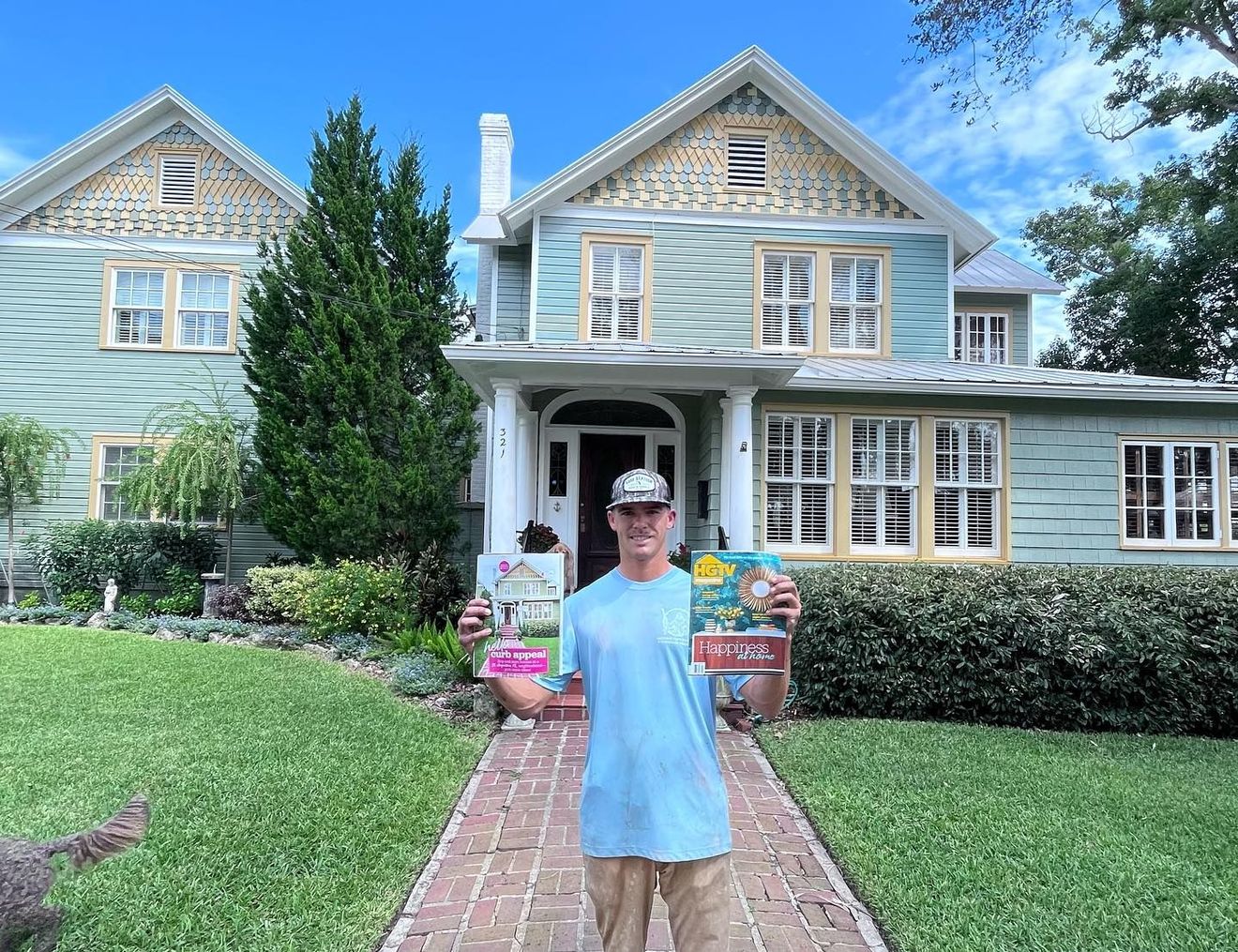 Man in front of a blue house with a book and plants. Sunny day, green grass, light blue shirt, white hat.