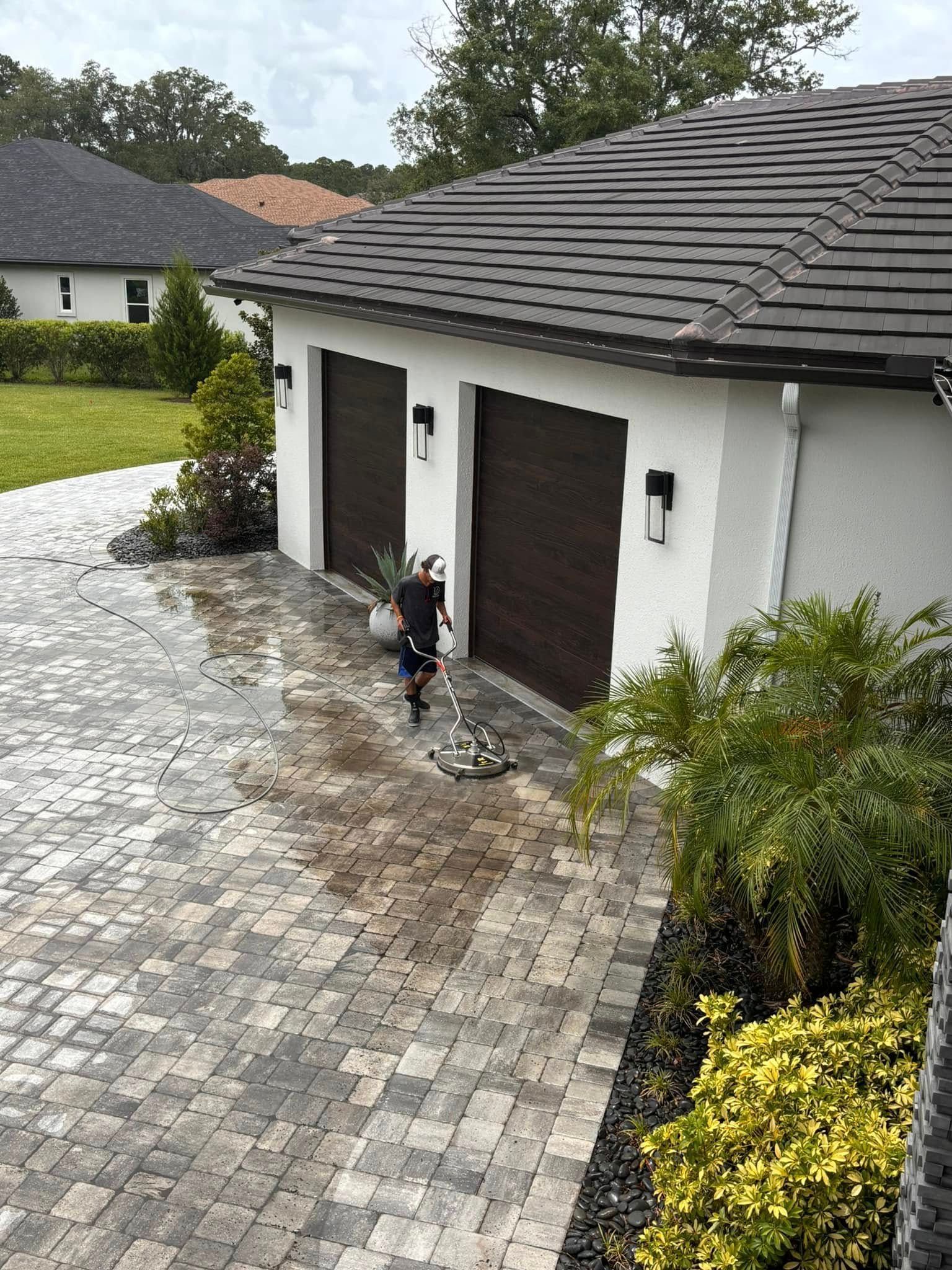 Person pressure washing a brick driveway in front of a two-car garage.